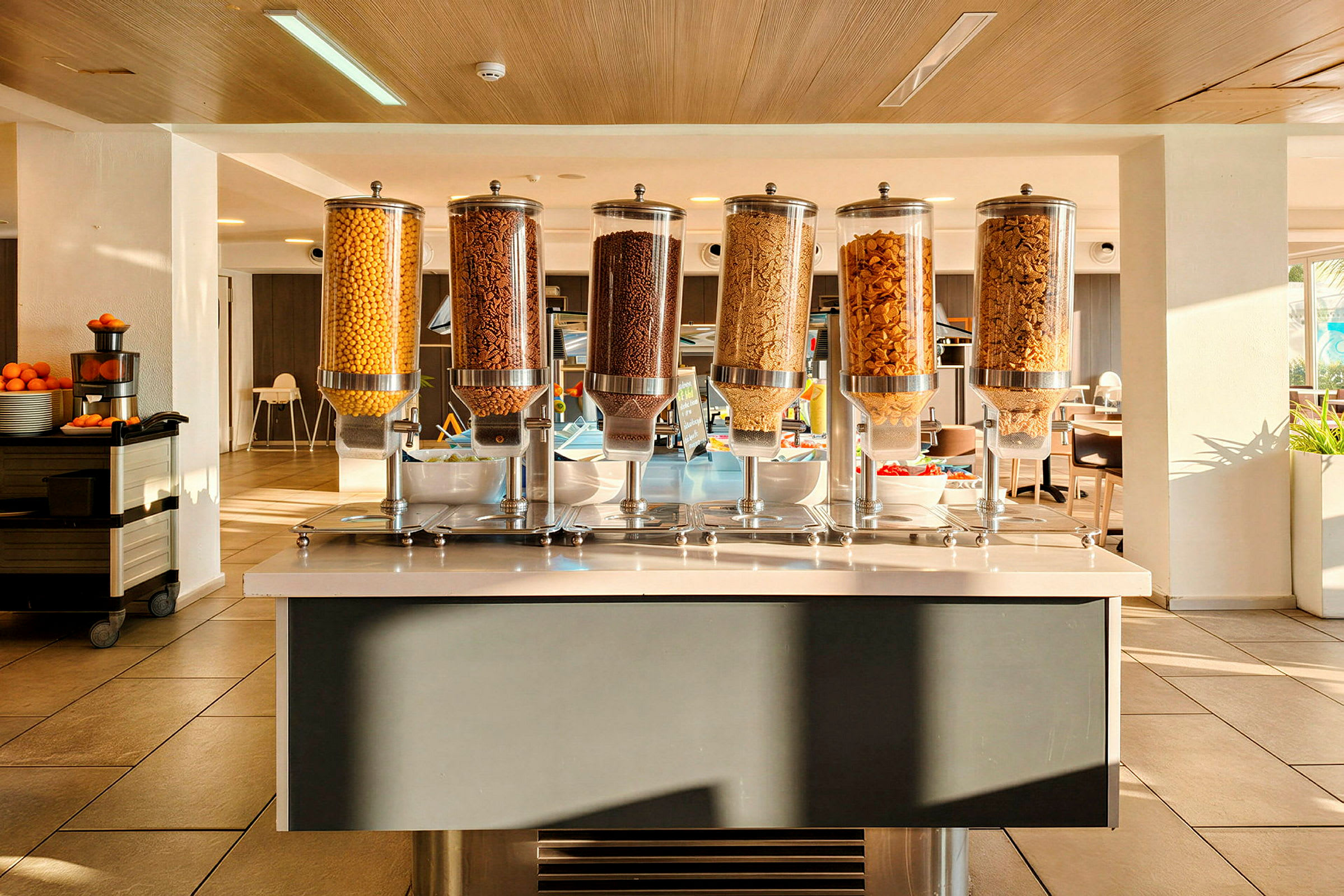 En morgenmadsbuffet med forskellige morgenmadsprodukter i dispensere og en frugtkurv i baggrunden. (AI-genereret tekst)