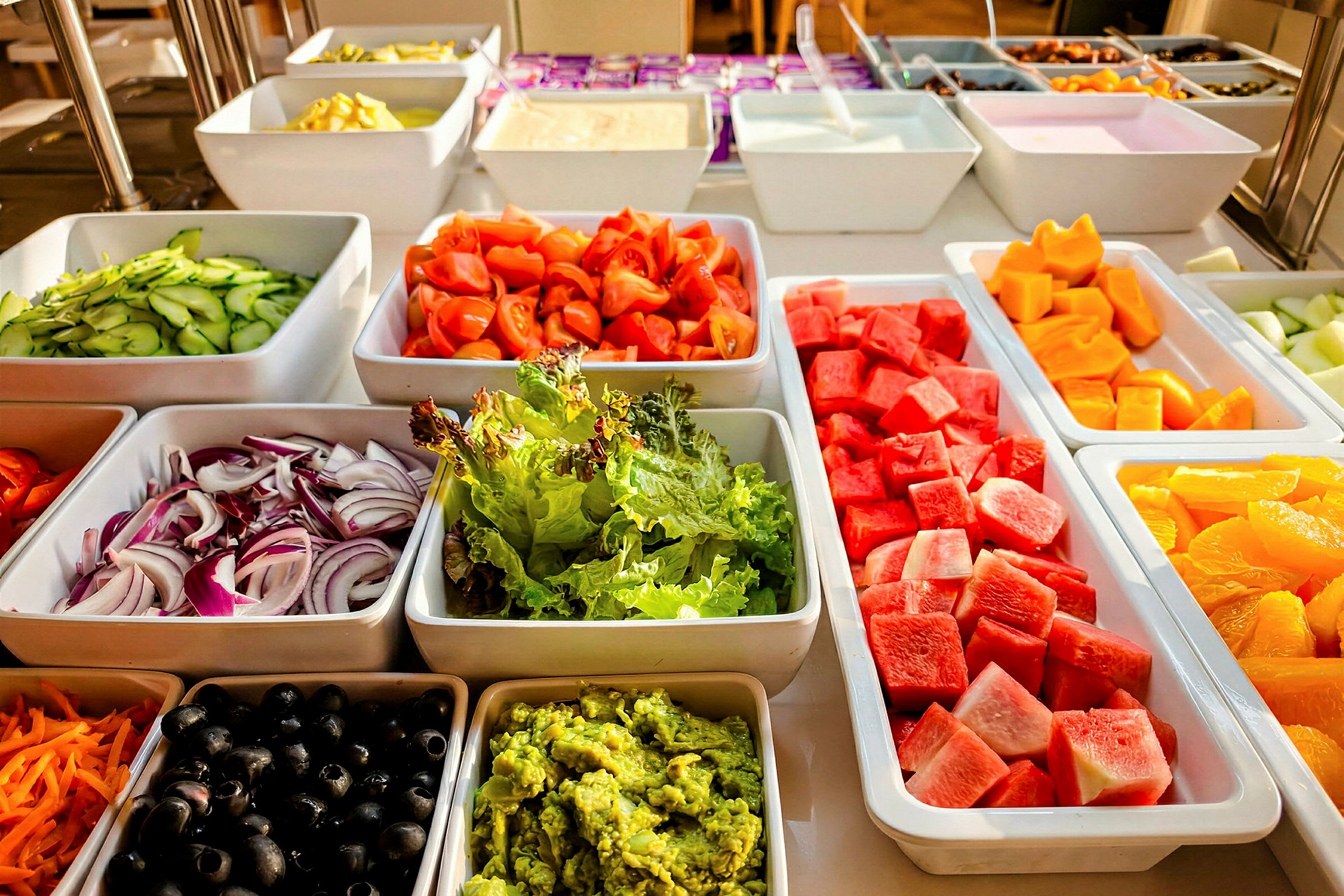 Et buffetbord med skåle med forskellige grøntsager og frugter, herunder salat, tomater, vandmelon og appelsiner. (AI-genereret tekst)
