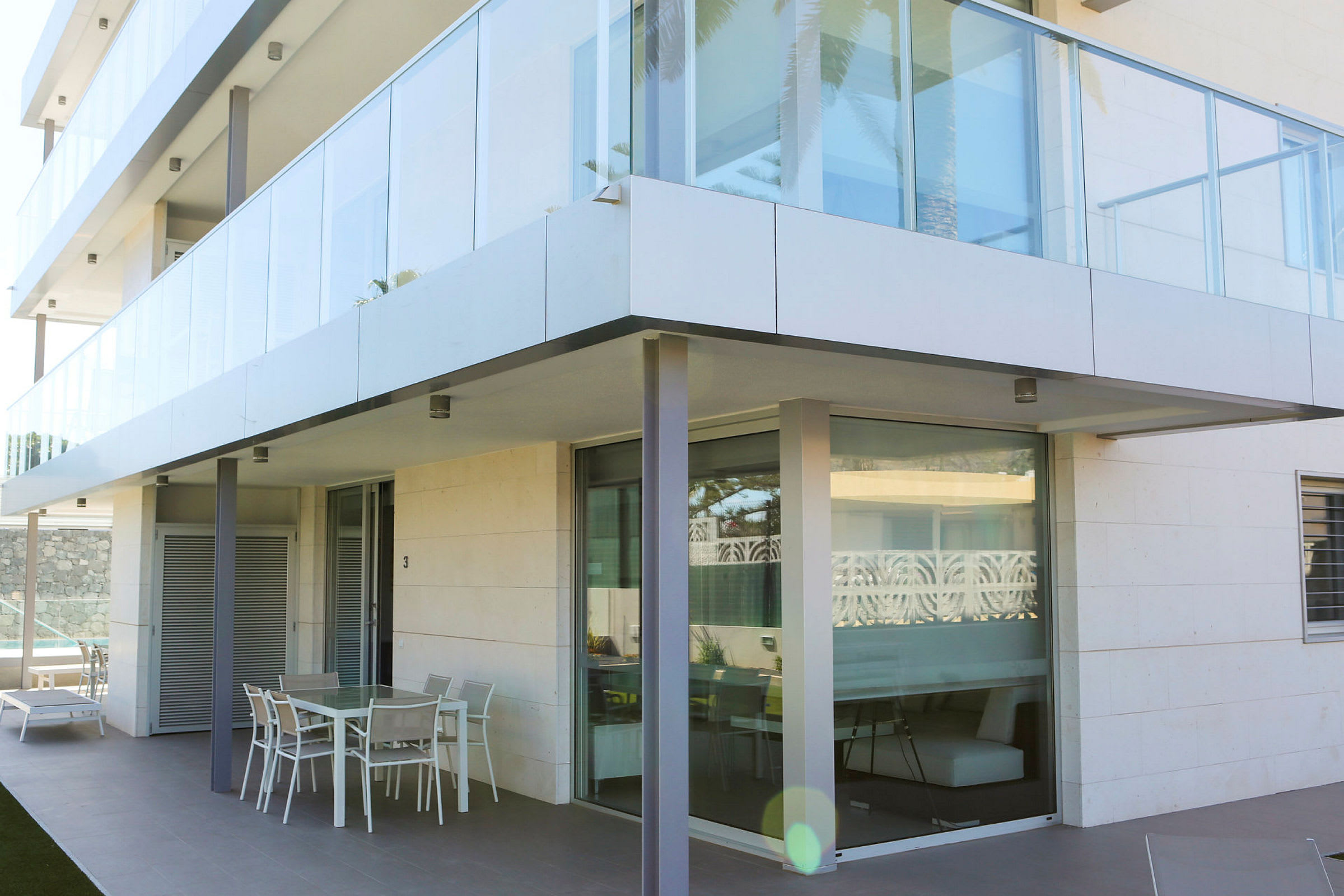 Det moderne flerfamiliehus har en glasfacade med en udendørs terrasse på stueetagen, der inkluderer havemøbler. (AI-genereret tekst)