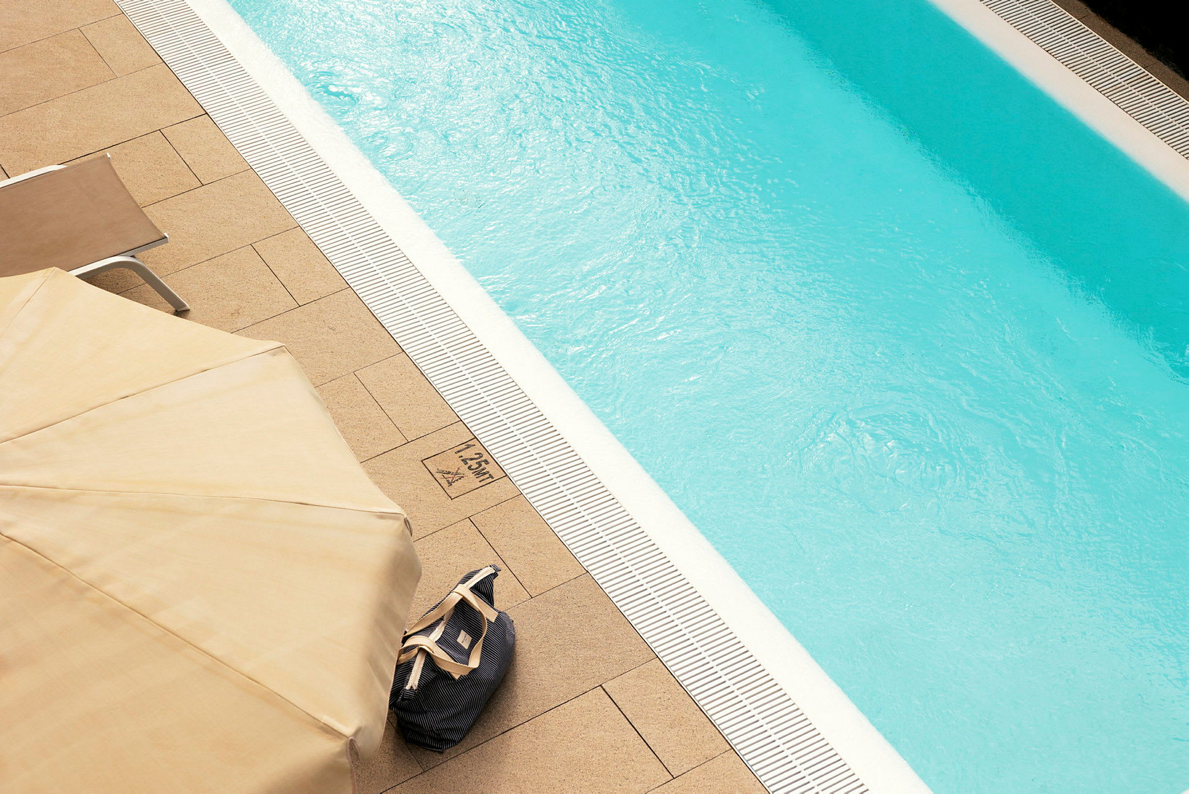 Billede af en pool med klart vand, omgivet af terrassefliser. En liggestol, parasol og taske ses ved kanten. (AI-genereret tekst)