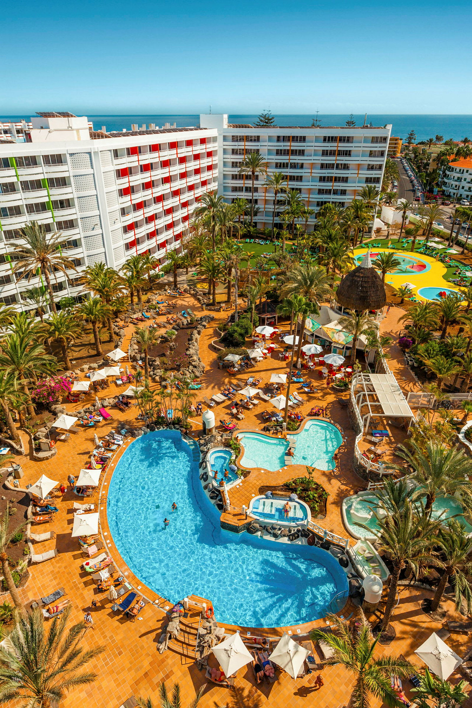 Udsigt over et hotelområde med hvide bygninger, swimmingpools og palmer. (AI-genereret tekst)
