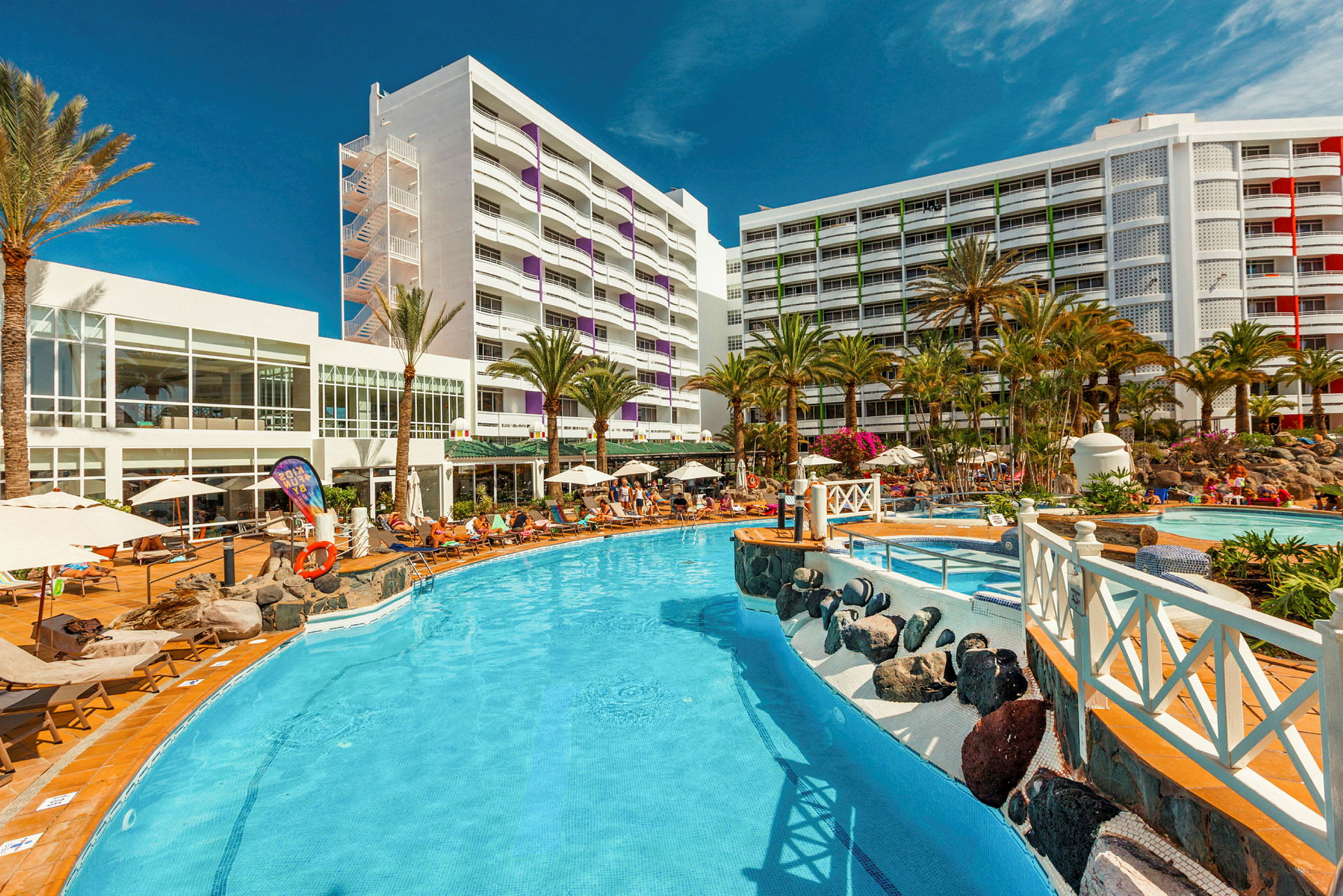 Billede af hotelområde med pool, omkringliggende solsenge og palmer. (AI-genereret tekst)