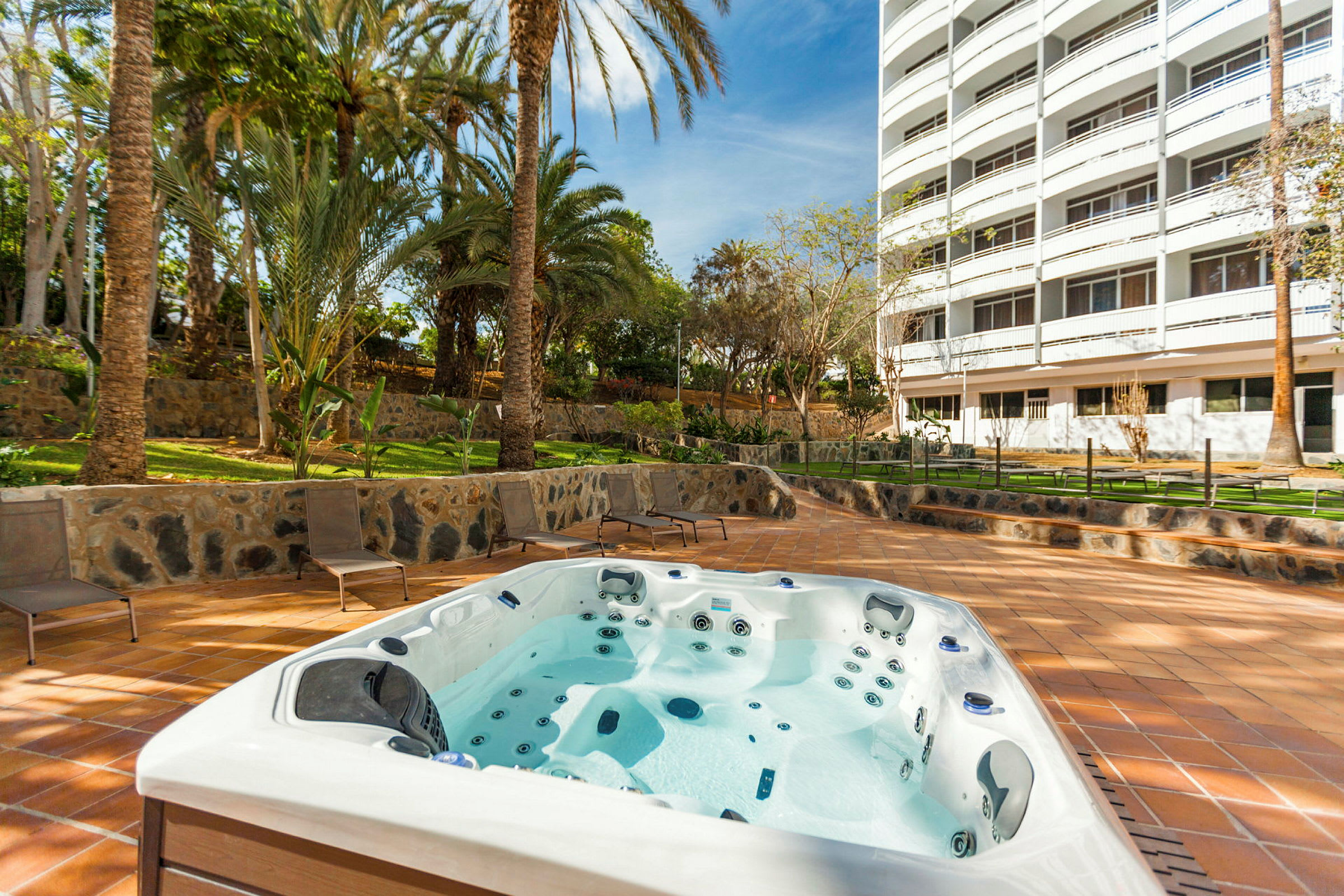 Billede af en udendørs jacuzzi beliggende tæt på en fleretagers bygning, omgivet af grønne områder og palmer. (AI-genereret tekst)