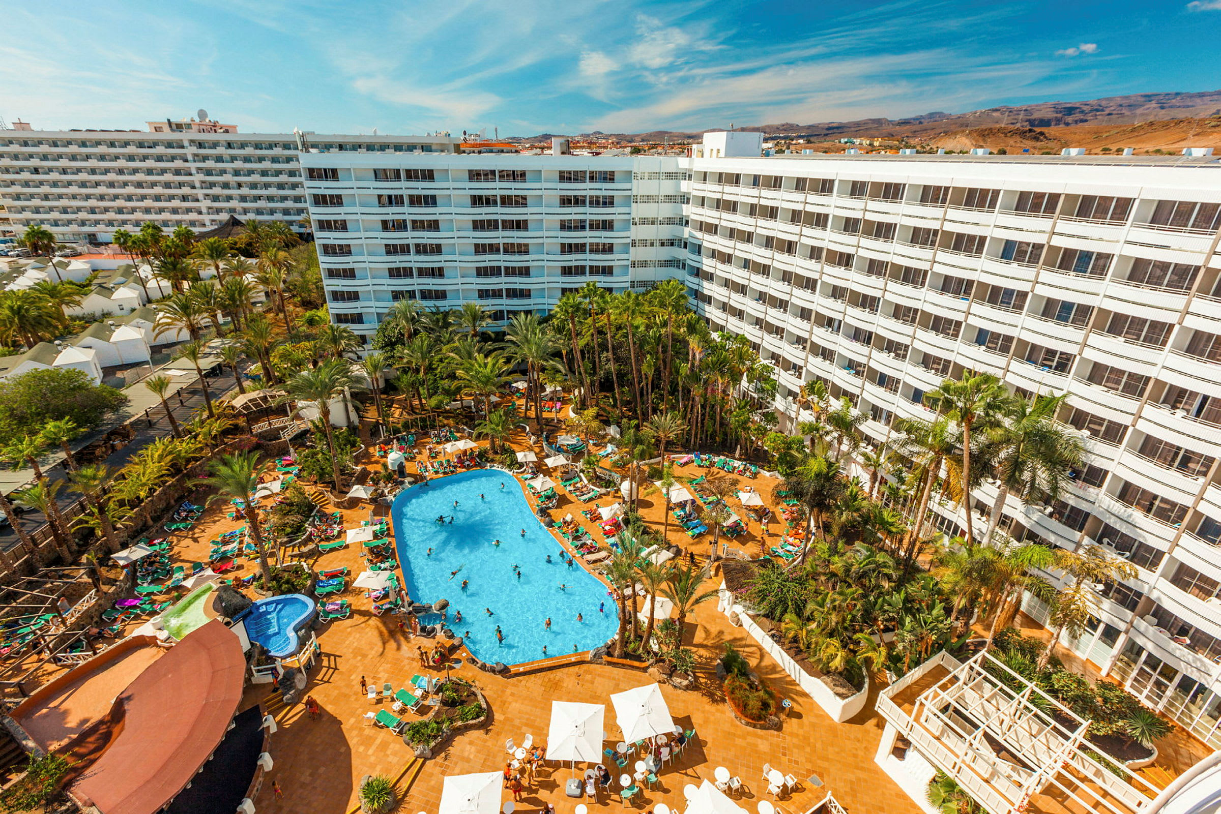 Hotelområde med stor bygning, palmer og swimmingpool. (AI-genereret tekst)