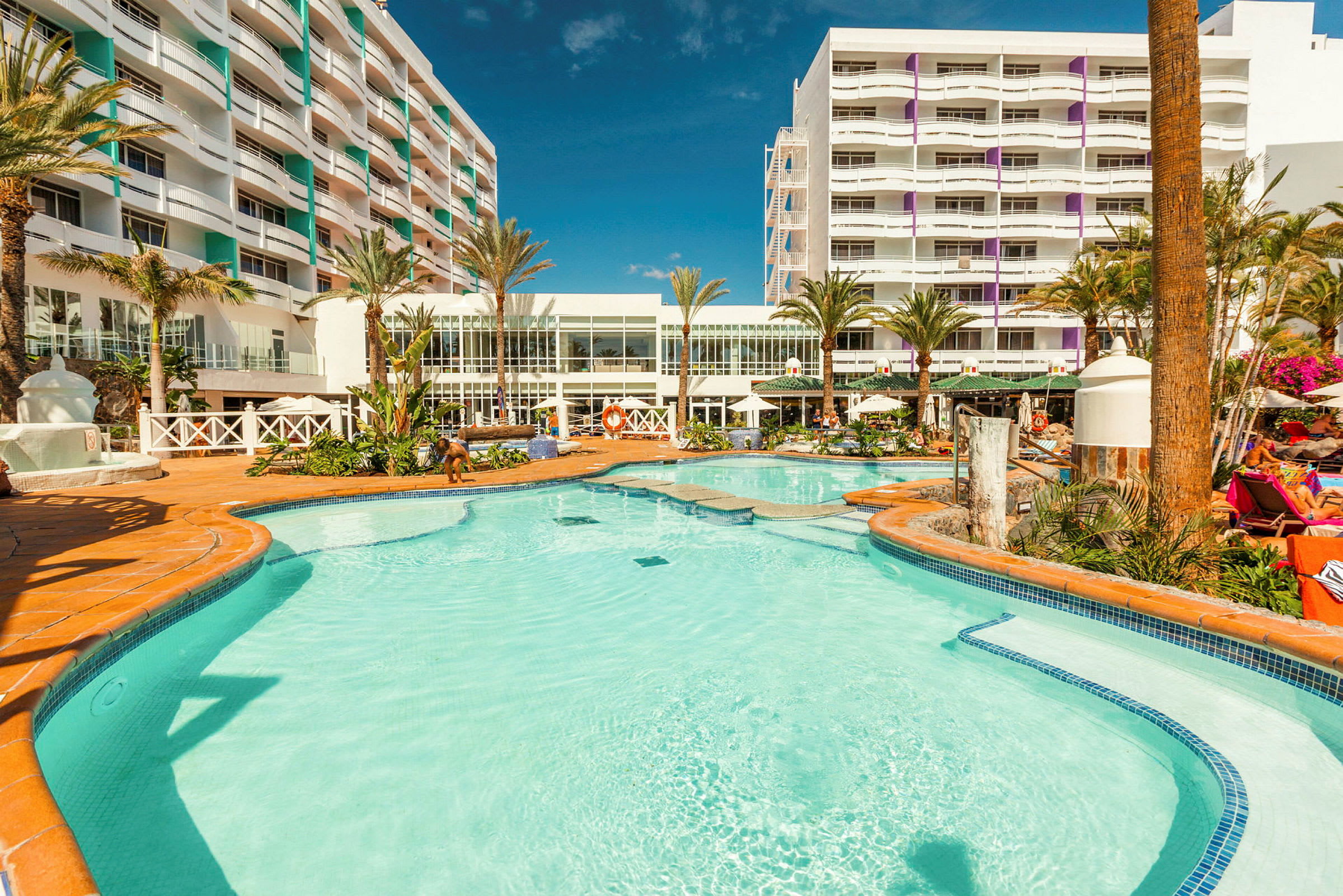 Udendørs swimmingpool med liggestole omkring og hotelbygning i baggrunden. (AI-genereret tekst)