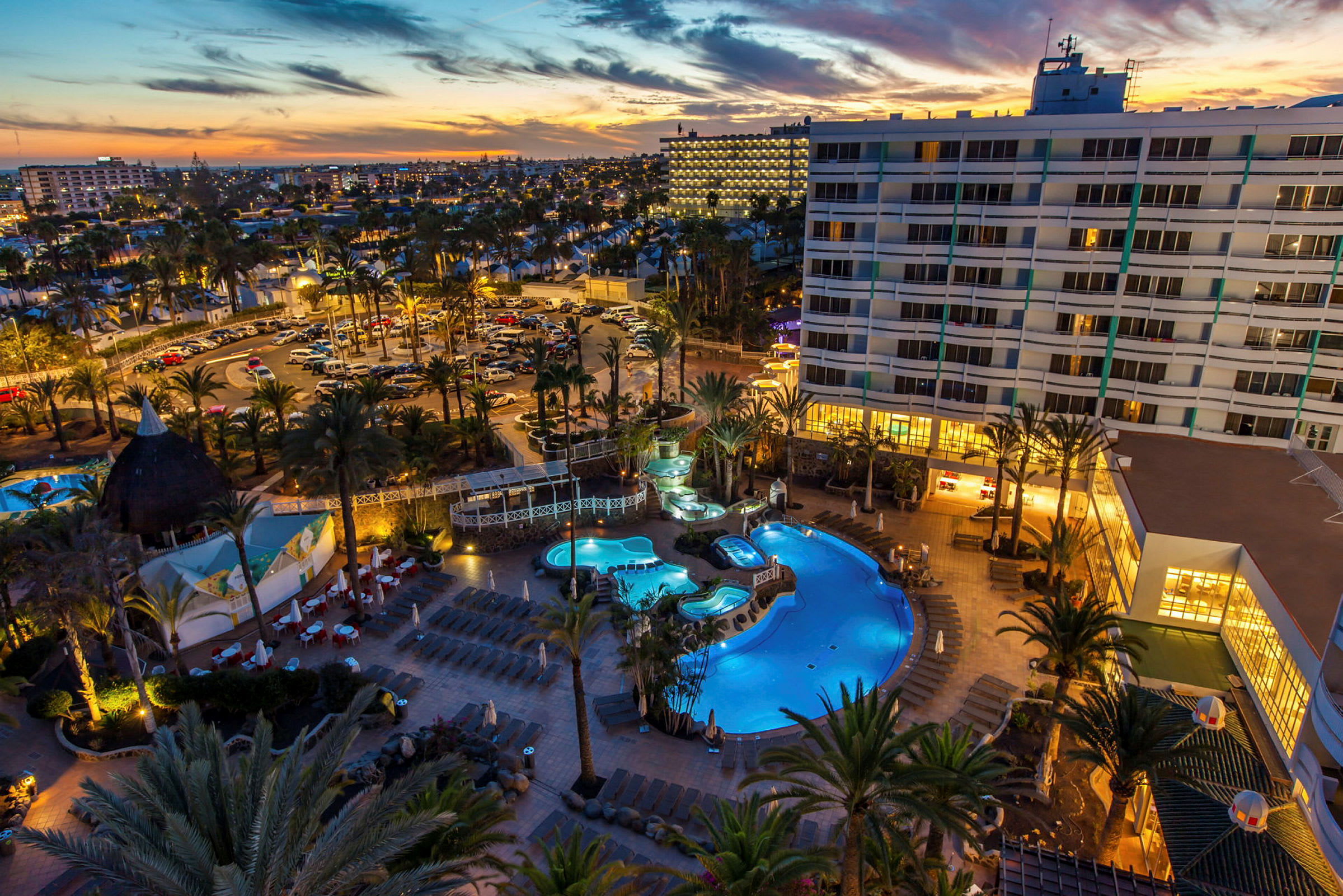 Billede af hotelområde med swimmingpools og omkringliggende bygninger ved solnedgang. (AI-genereret tekst)