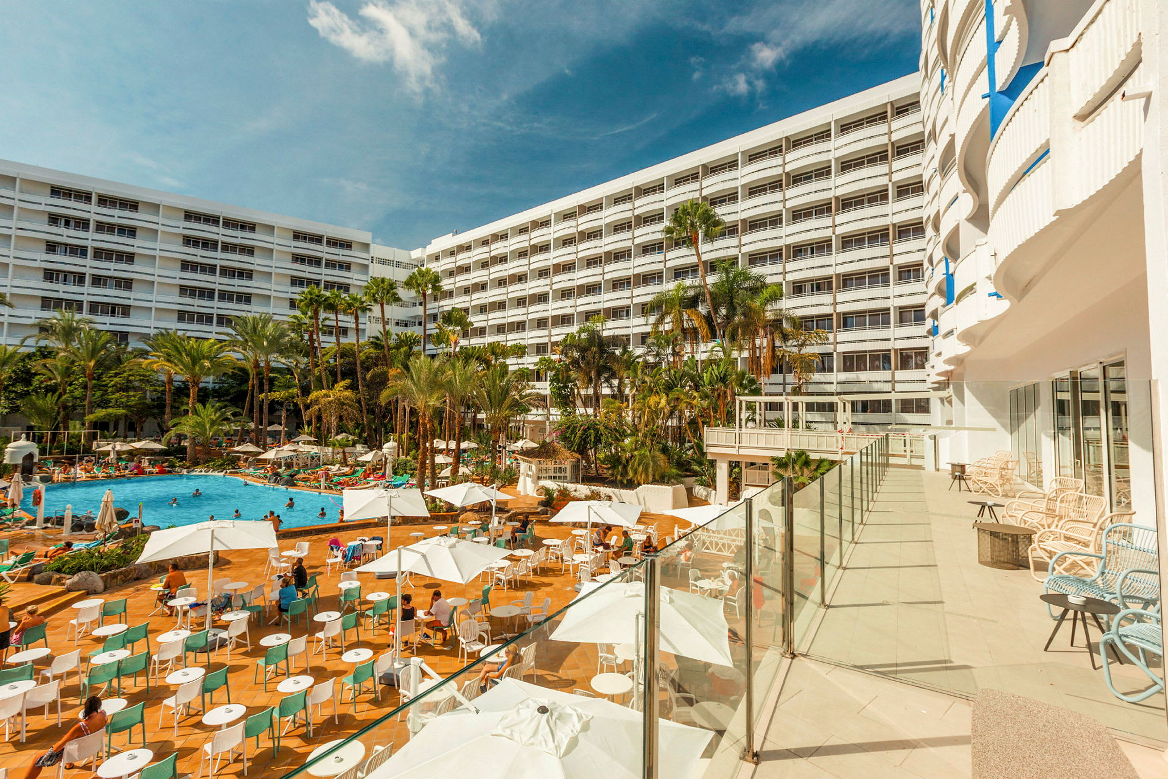 En hotelbygning med en stor udendørs pool omgivet af liggestole og parasoller. (AI-genereret tekst)