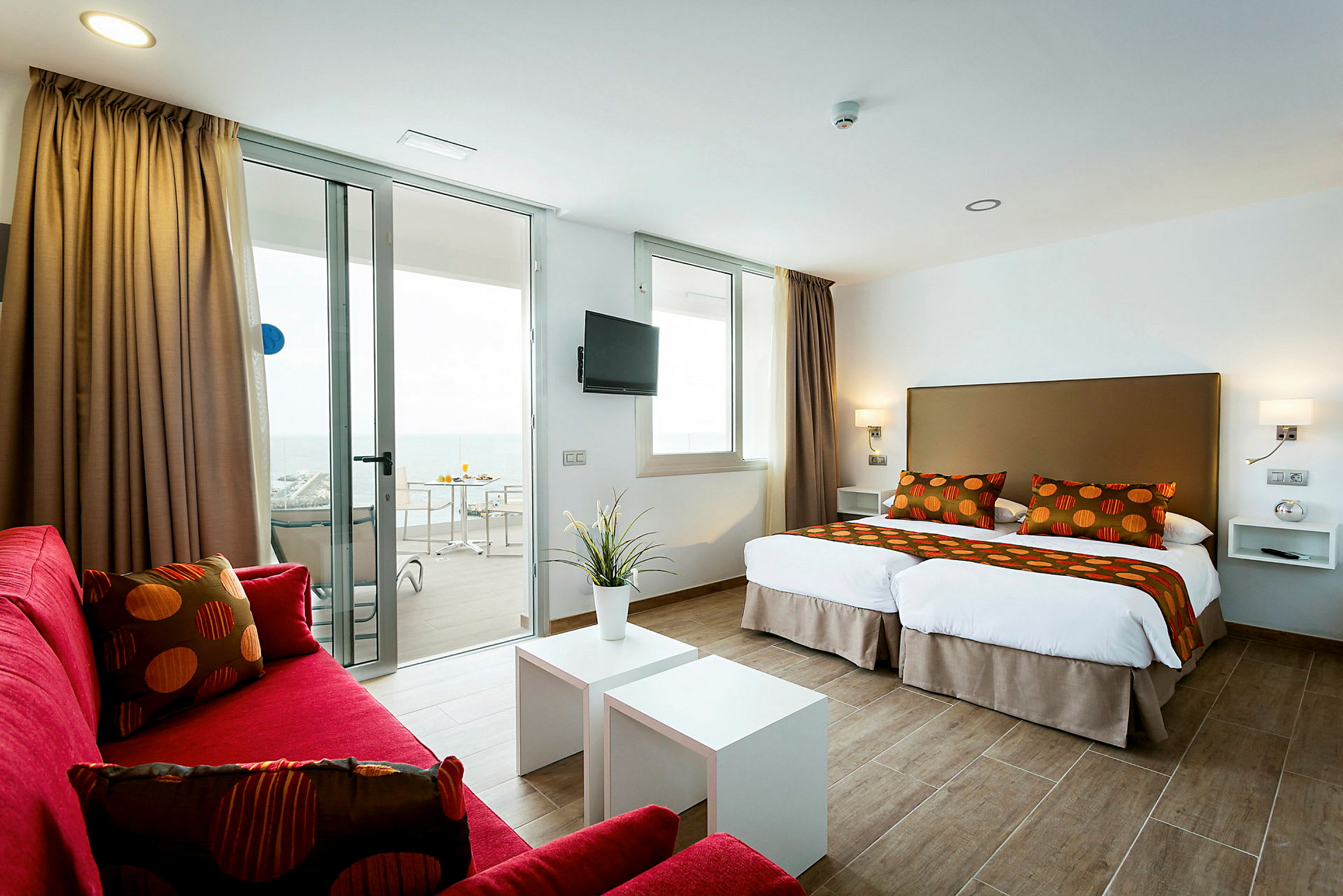 Et moderne hotelværelse med to enkeltsenge, røde sofaer og en altan med havudsigt. (AI-genereret tekst)