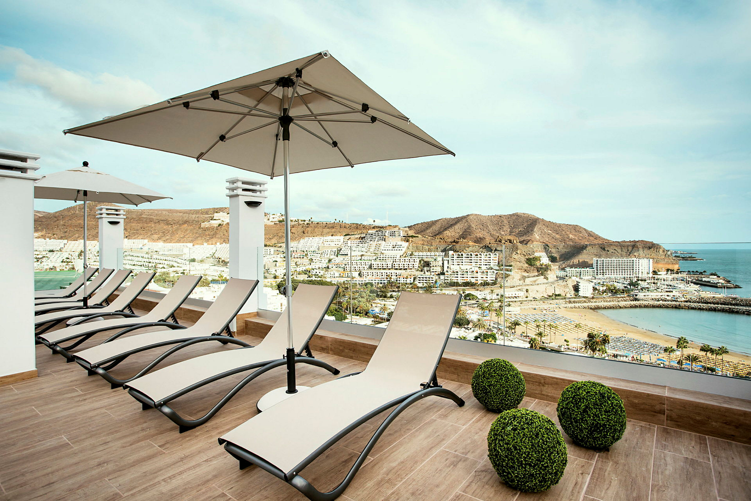Liggestole på en terrasse med udsigt over en strand og omkringliggende bygninger. (AI-genereret tekst)