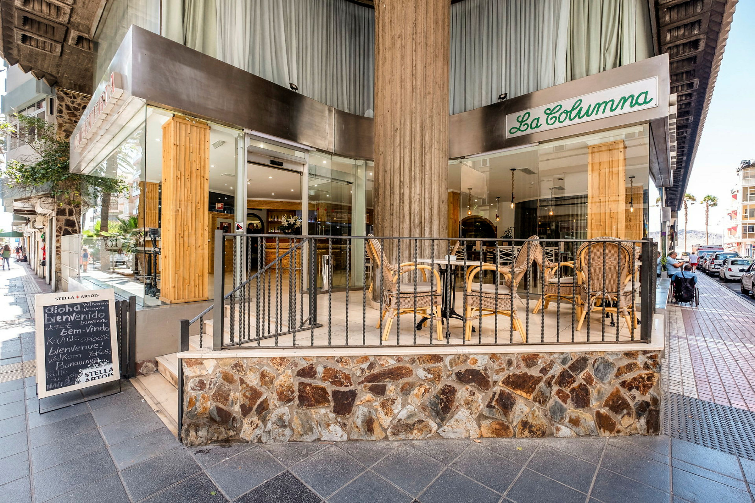 Indgangen til en restaurant med udendørs servering, hvor der står borde og stole. Restauranten hedder "La Columna" og ligger ved et fortov. (AI-genereret tekst)