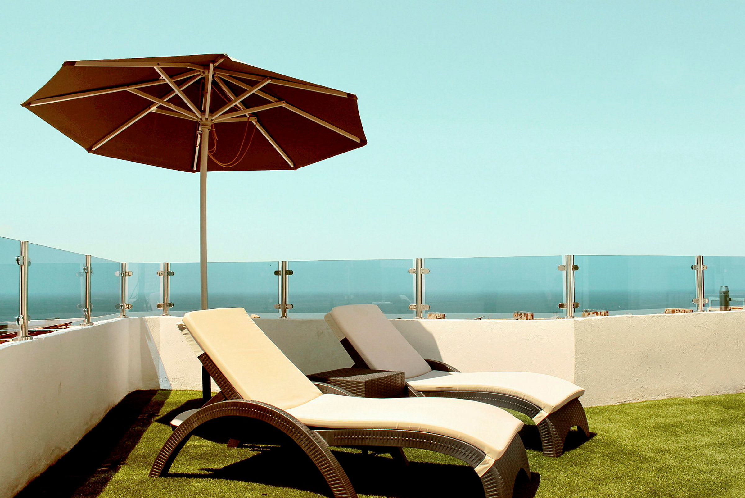 To liggestole med beige hynder og en brun parasol på en terrasse med glasrækværk og havudsigt. (AI-genereret tekst)