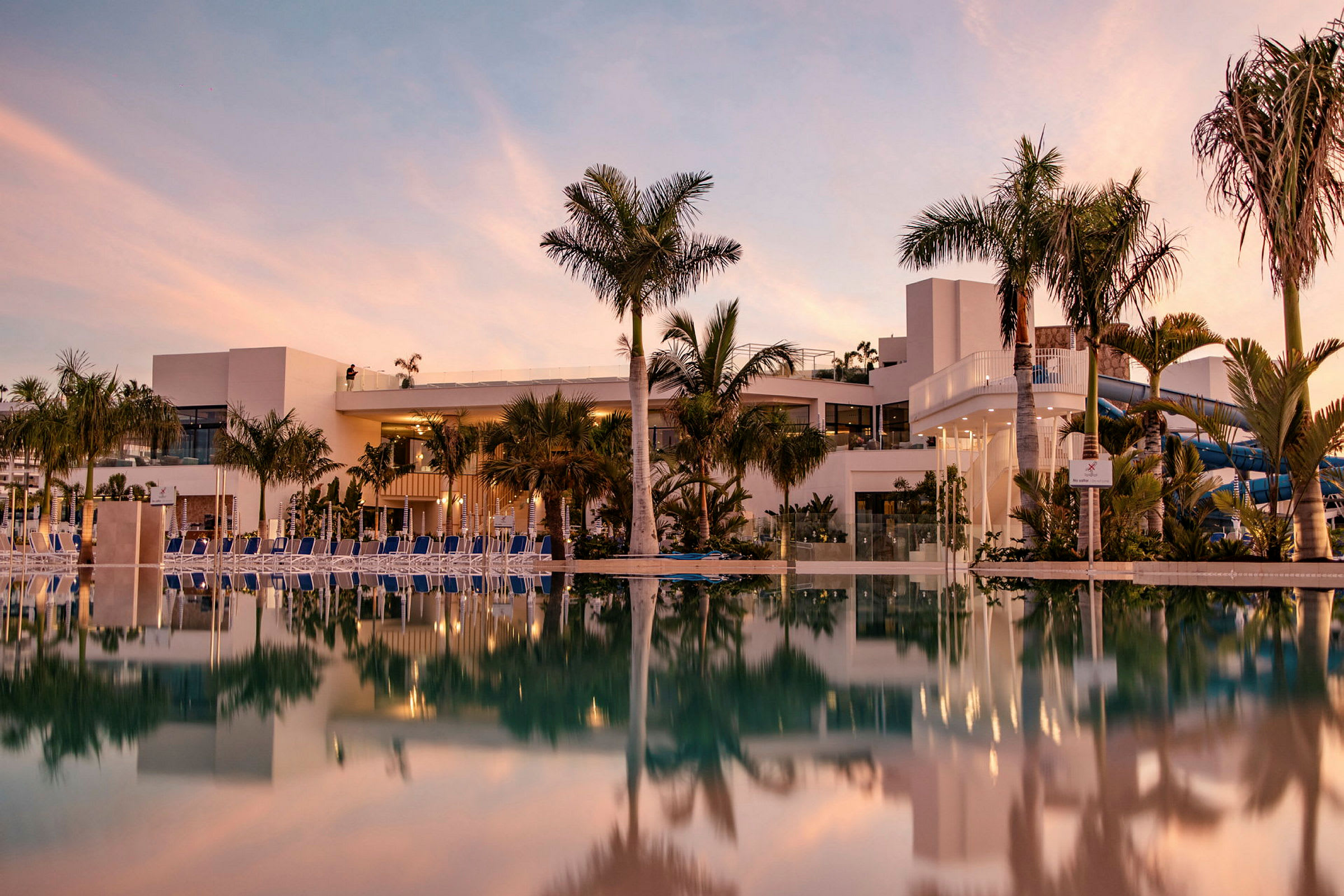 Hotelbygning og swimmingpool med spejlbillede ved solnedgang. (AI-genereret tekst)