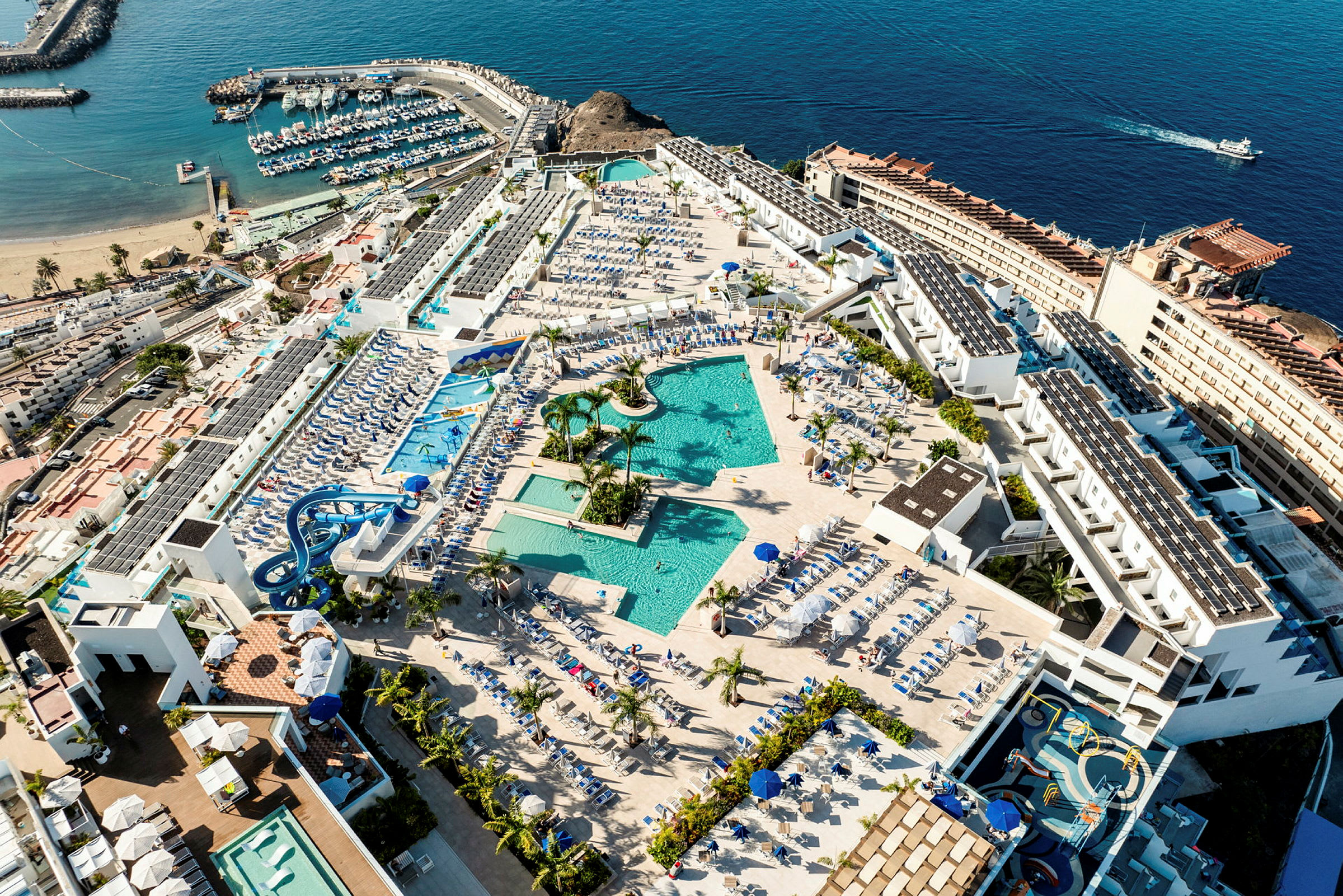 Luftfoto af hotelområde med pools, liggestole og omkringliggende bygninger tæt på havet. (AI-genereret tekst)