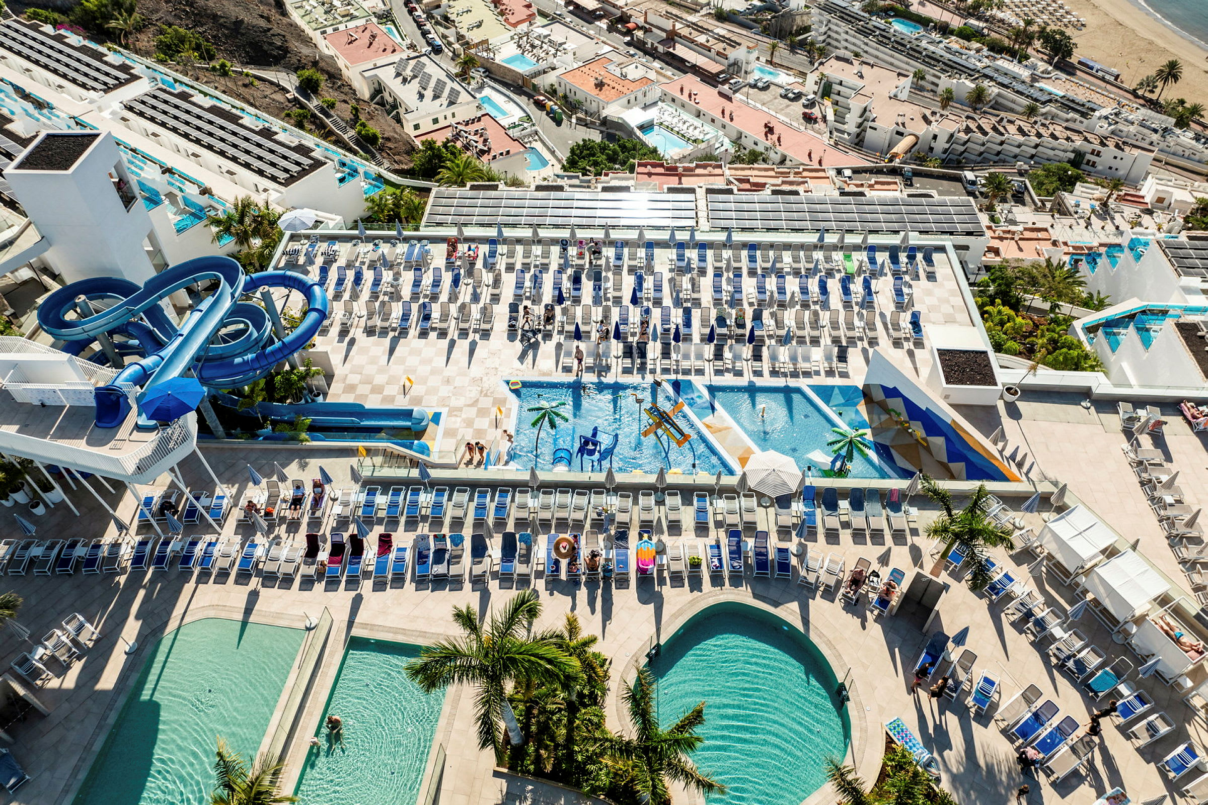 Luftfoto af en resort med flere pools, liggestole og vandrutsjebaner. (AI-genereret tekst)