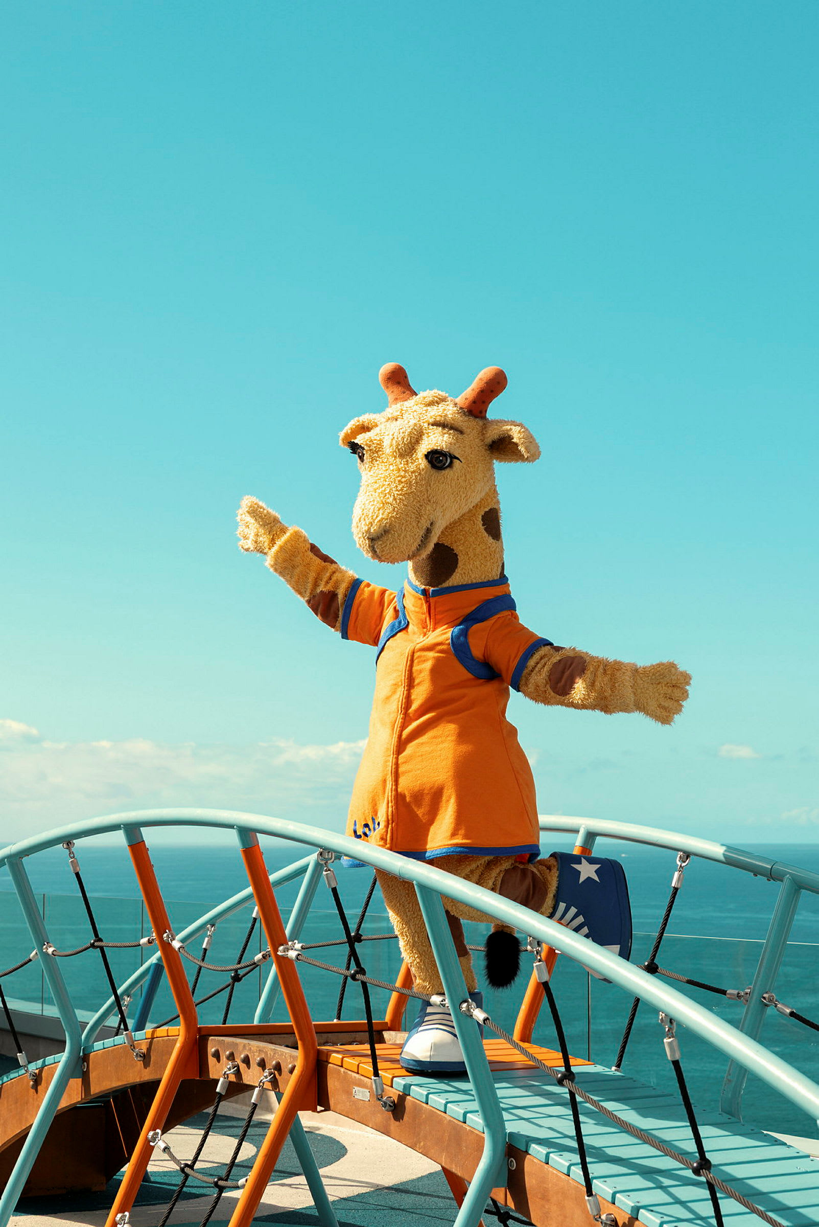 En person klædt som en girafmaskot står på en legeplads med havet i baggrunden. (AI-genereret tekst)