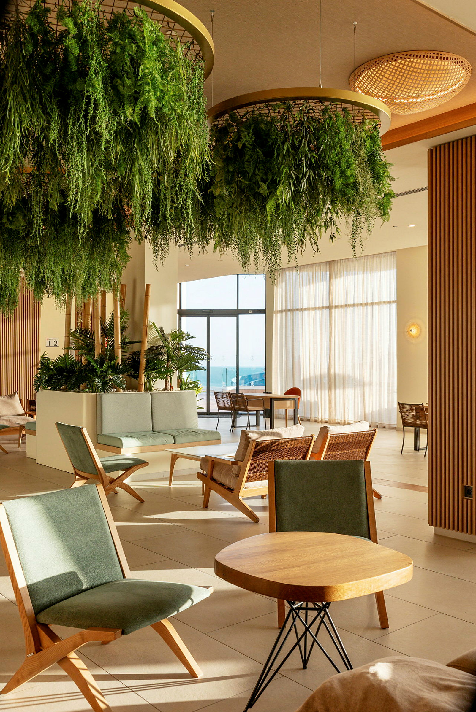 En hotellets lobby med grønne planter hængende fra loftet, siddegrupper med træstole og borde, og glasdøre der fører ud til en solrig terrasse. (AI-genereret tekst)