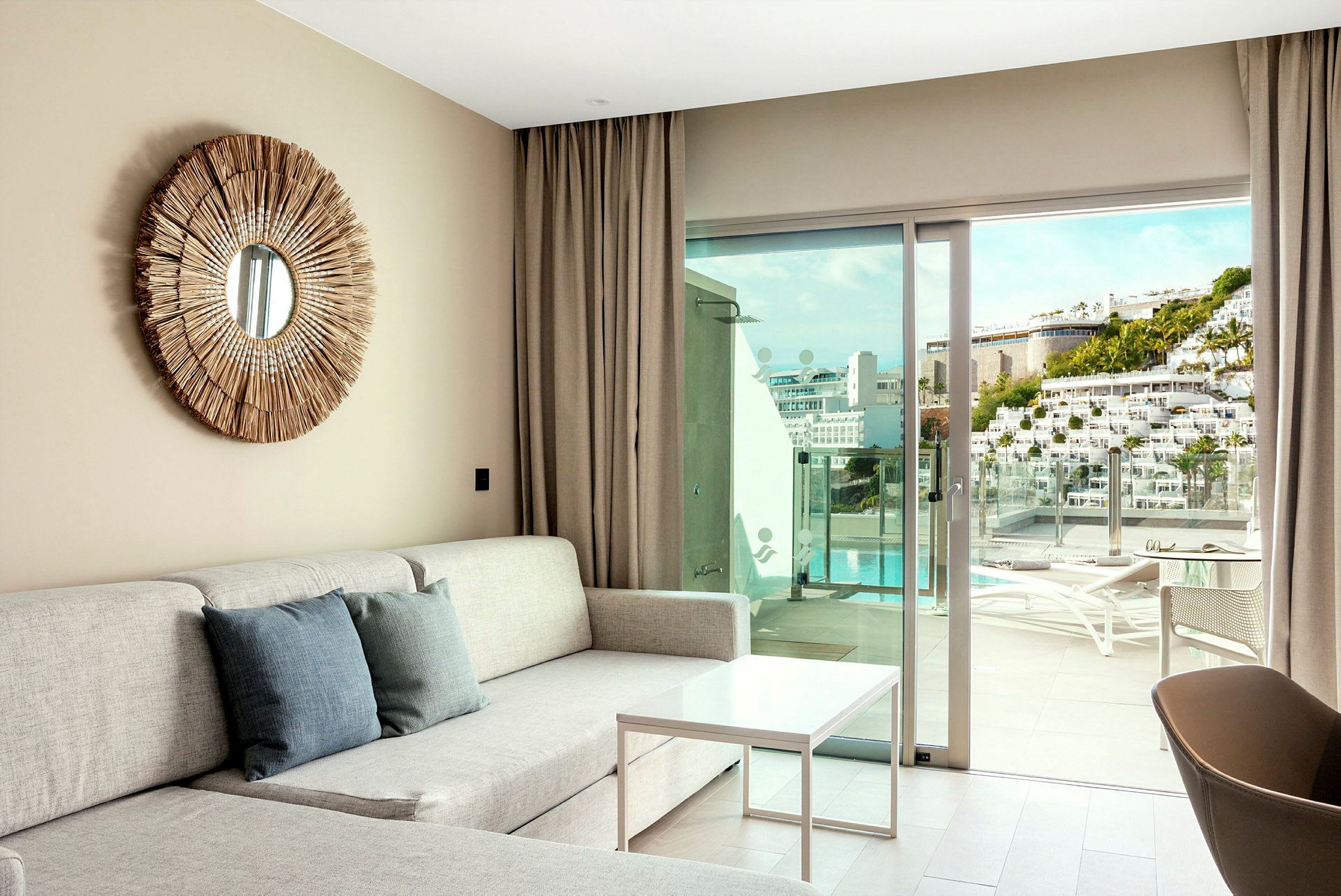 En moderne dagligstue med sofa, to pyntepuder og en rund vægdekoration. Glasdøre åbner op til en solrig altan med udsigt til pool. (AI-genereret tekst)