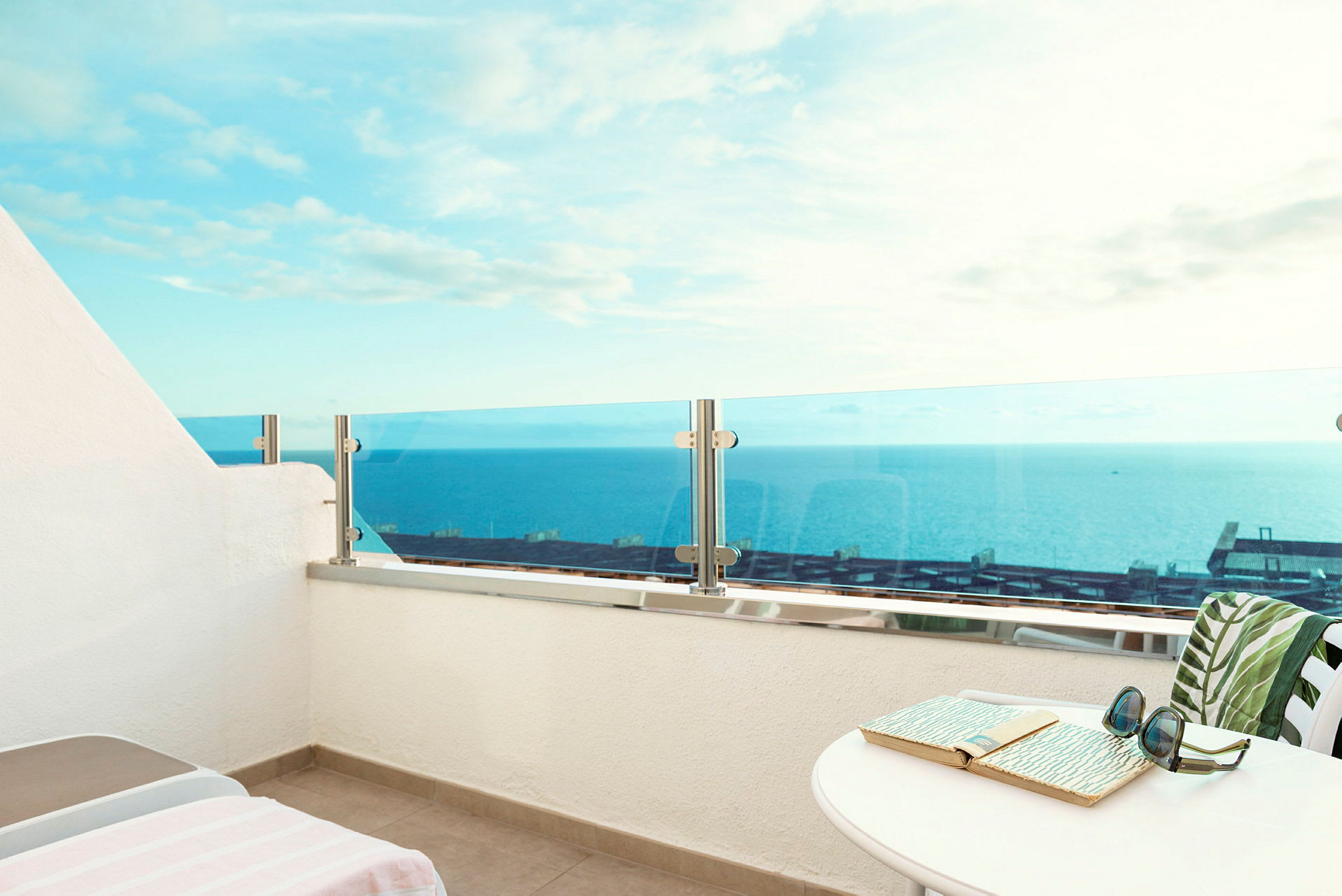 Udsigt over havet fra en balkon med glasrækværk, en solseng og et bord med en bog og solbriller. (AI-genereret tekst)