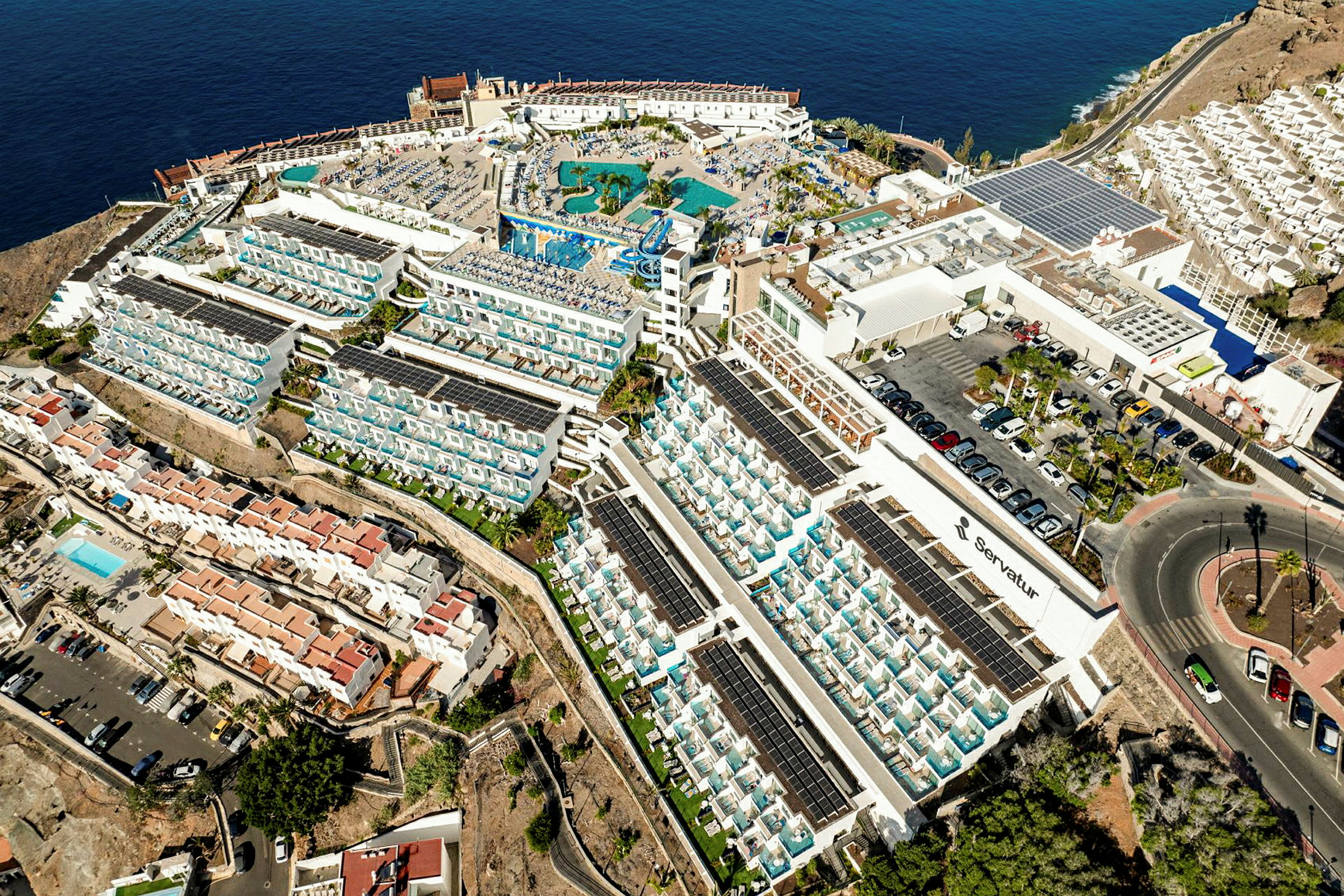 Luftfoto af et stort hotelkompleks ved havet med poolområde og omkringliggende boliger. (AI-genereret tekst)