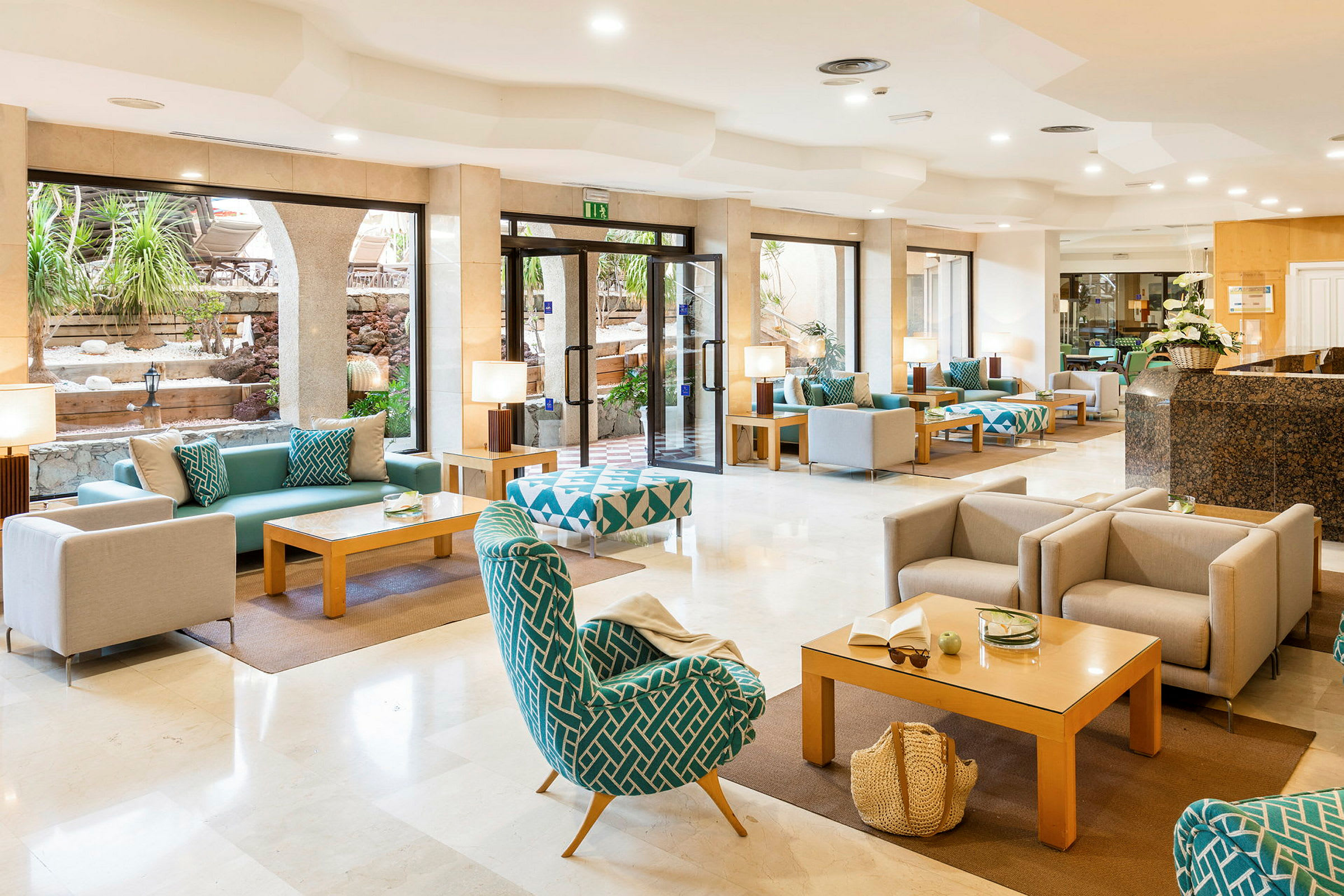 Hotellets lobby med siddepladser og sofagrupper samt store vinduer mod en udendørs gårdhave. (AI-genereret tekst)