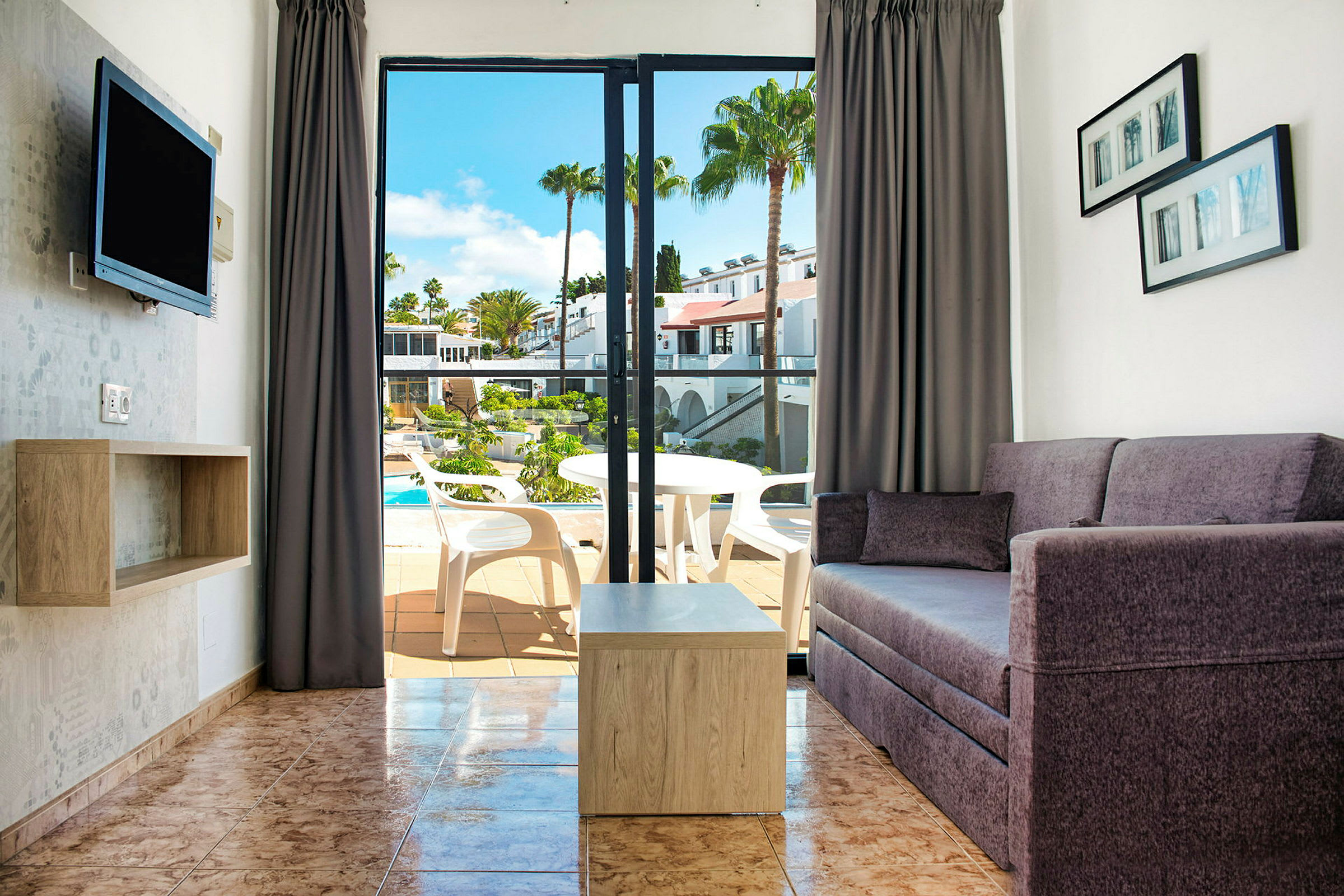 Udsigt over en stue med sofa og bord samt en åbning til en balkon med udsigt over en pool. (AI-genereret tekst)