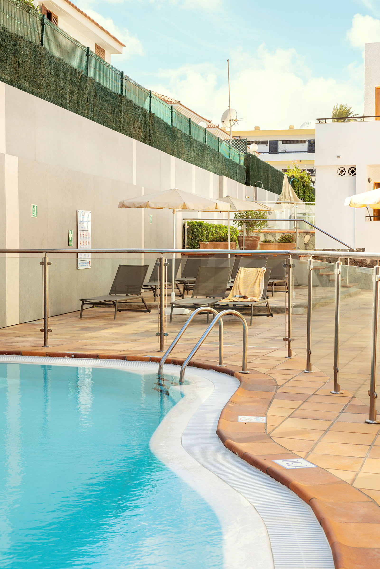 En udendørs swimmingpool med liggestole og parasoller i baggrunden. (AI-genereret tekst)