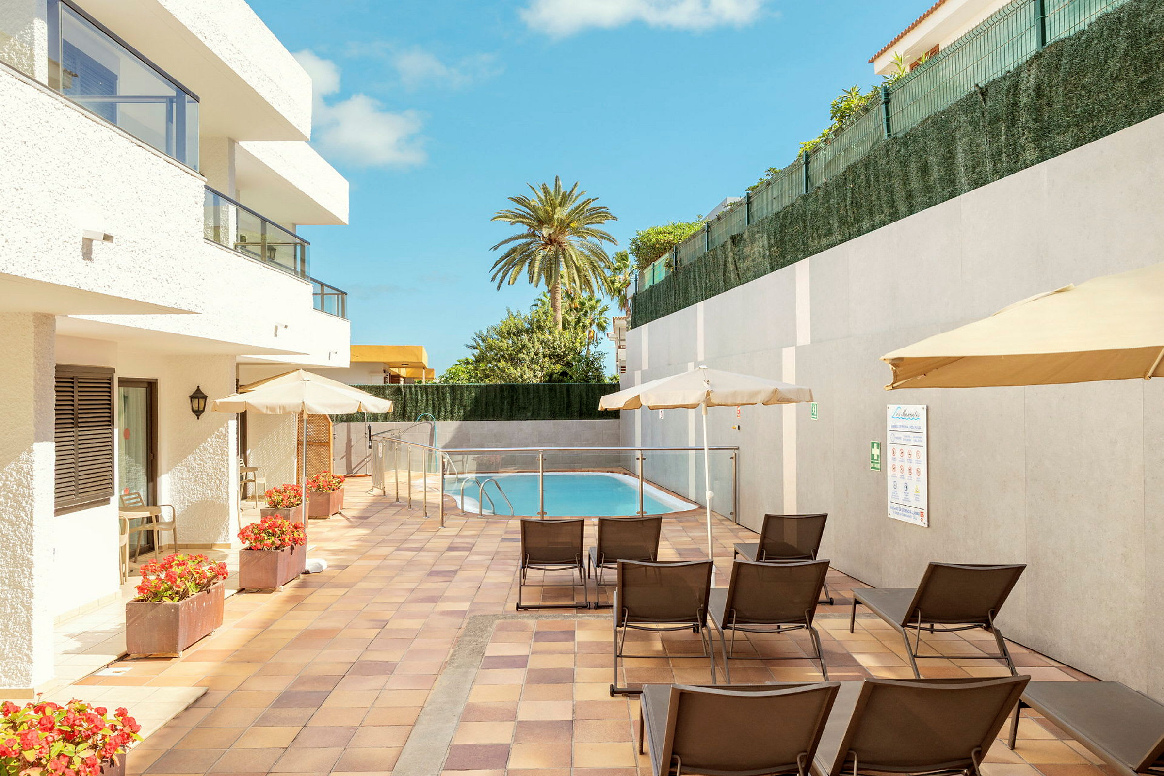 Et hotelområde med en lille pool, solsenge og parasoller omgivet af hvide bygninger og betonvægge. (AI-genereret tekst)