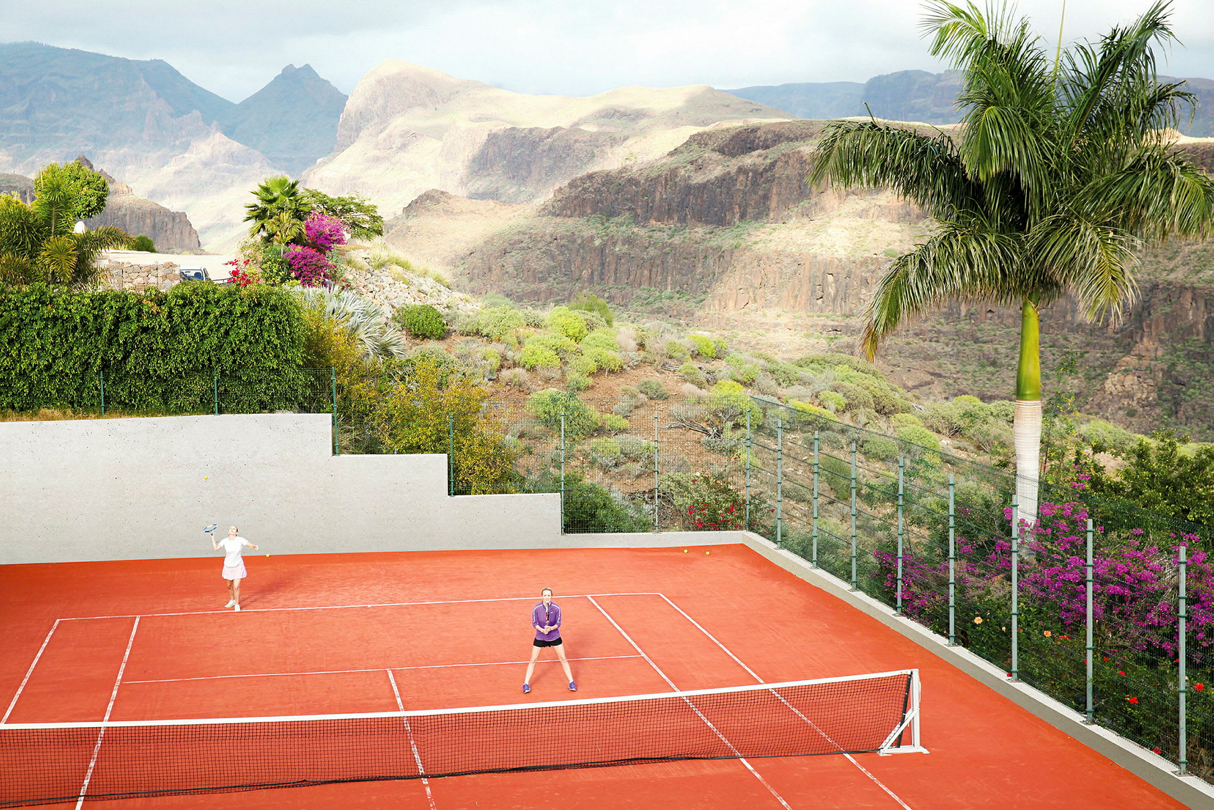 To personer spiller tennis på en rød tennisbane, omgivet af en grøn dal. (AI-genereret tekst)