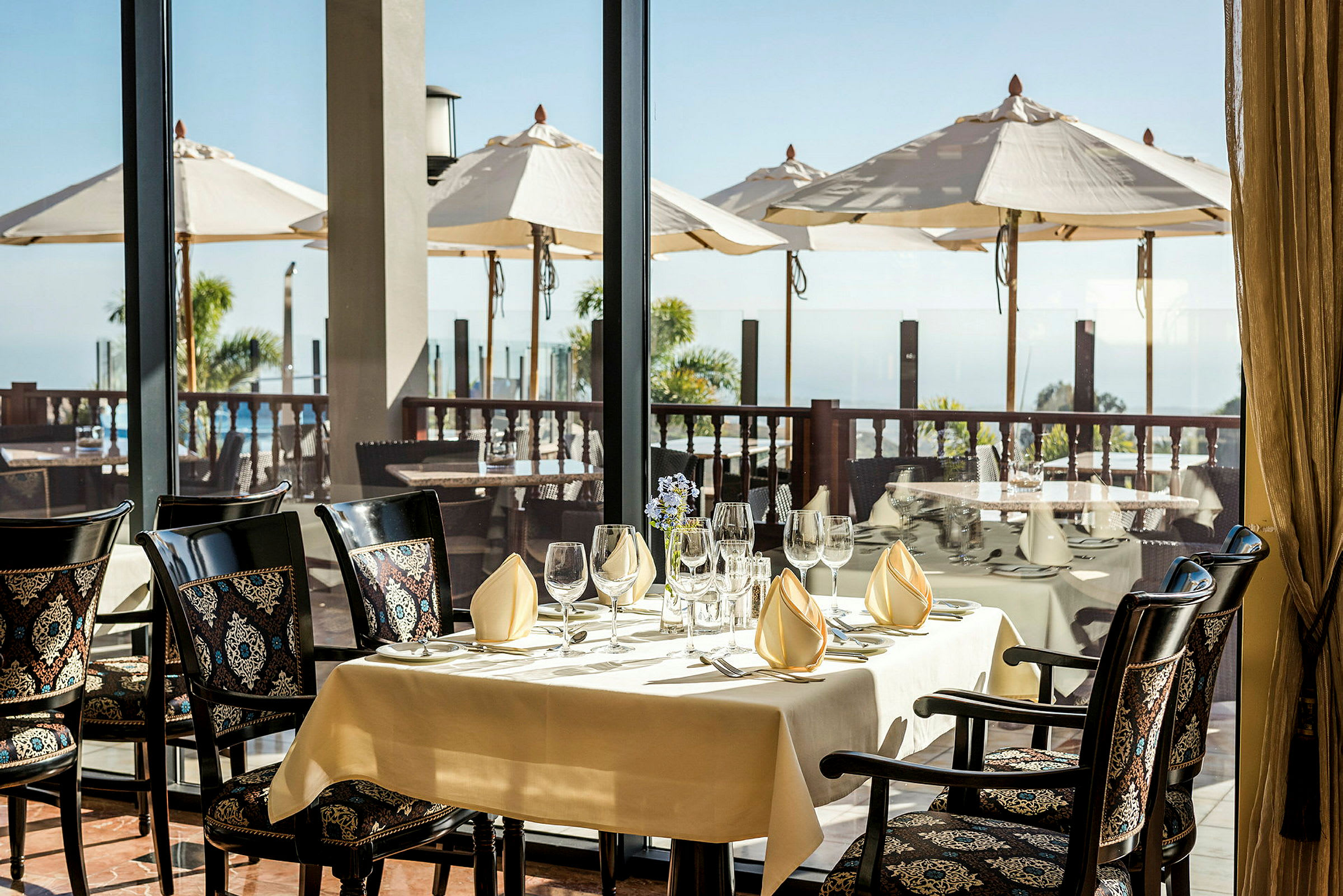 Et dækket bord i en restaurant med udsigt over en terrasse med parasoller og havet. (AI-genereret tekst)