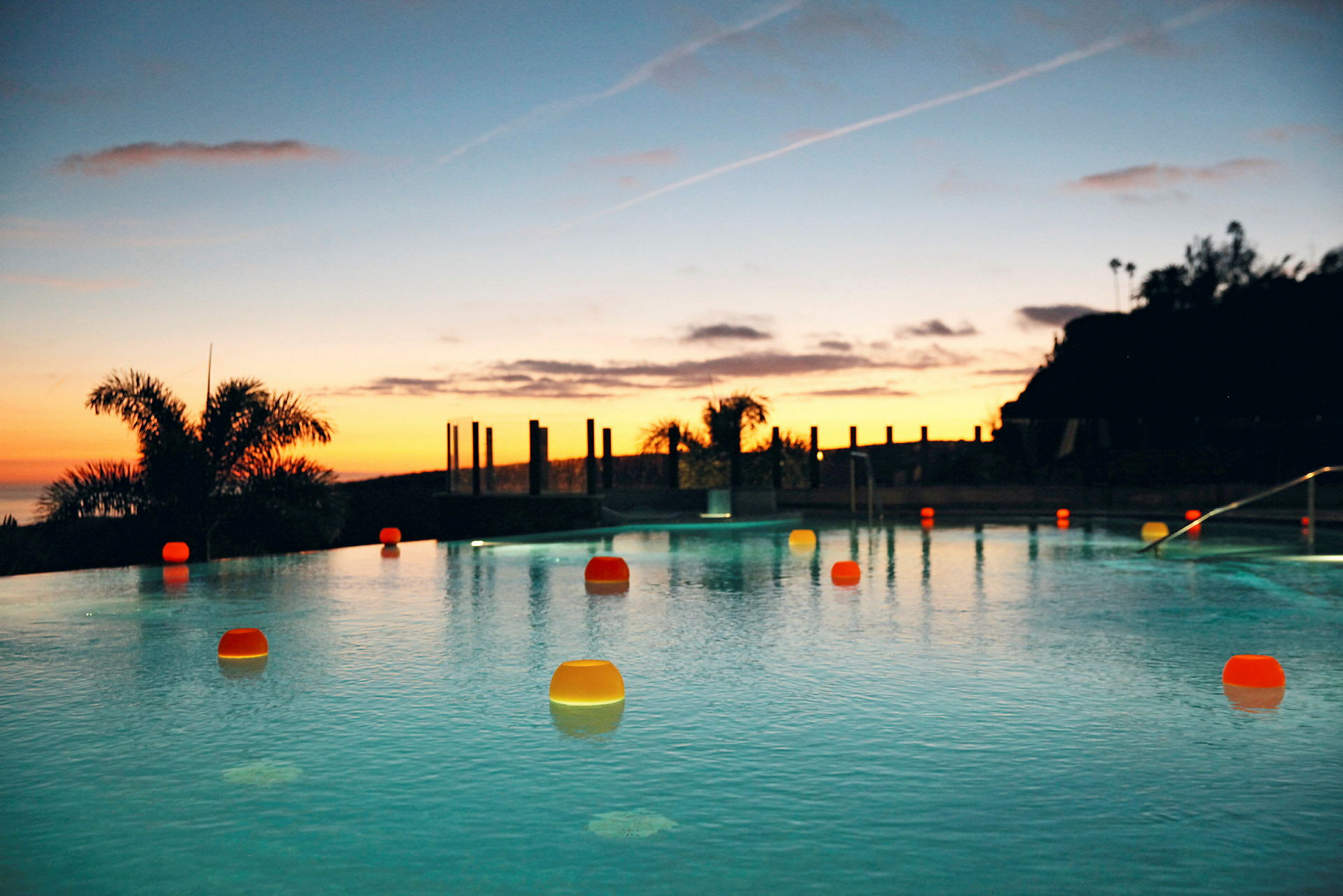 Aftenudsigt over en pool omgivet af palmer og orange belysning, med solnedgangen i baggrunden. (AI-genereret tekst)