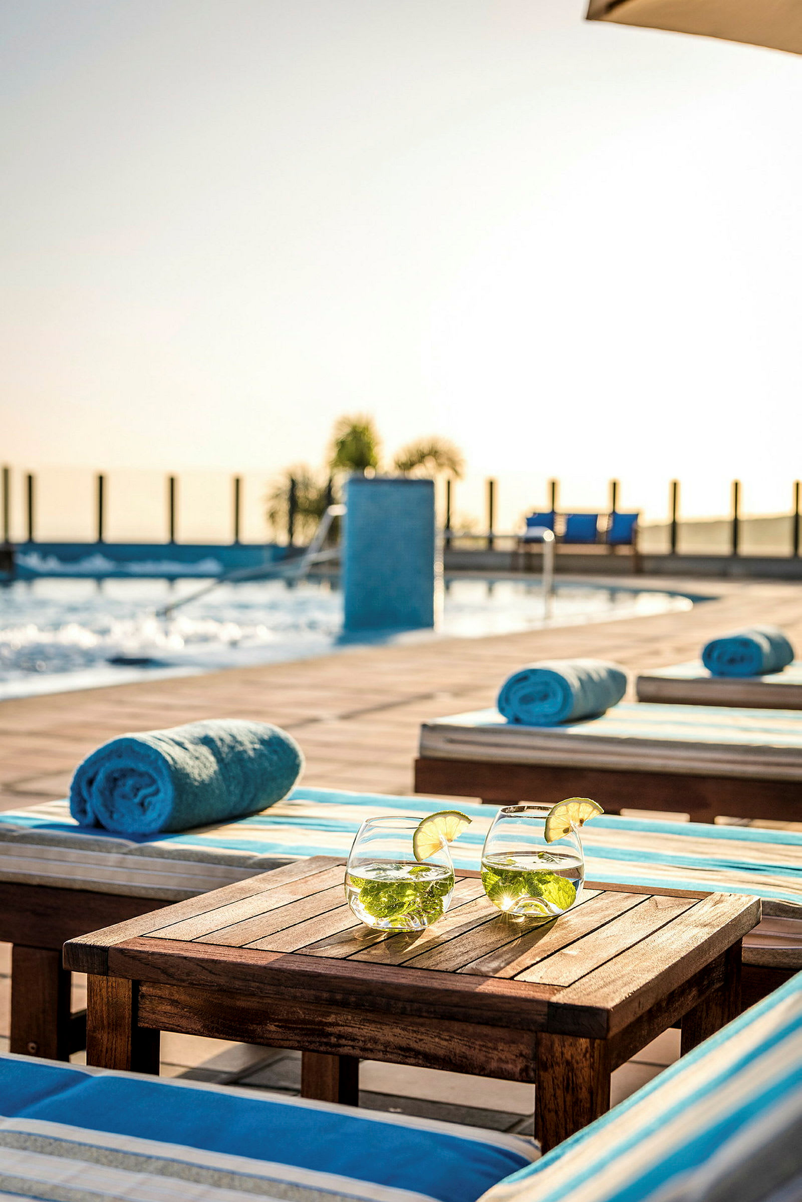 To glas med drikke, pyntet med lime, står på et bord ved liggestole og håndklæder nær en pool. (AI-genereret tekst)