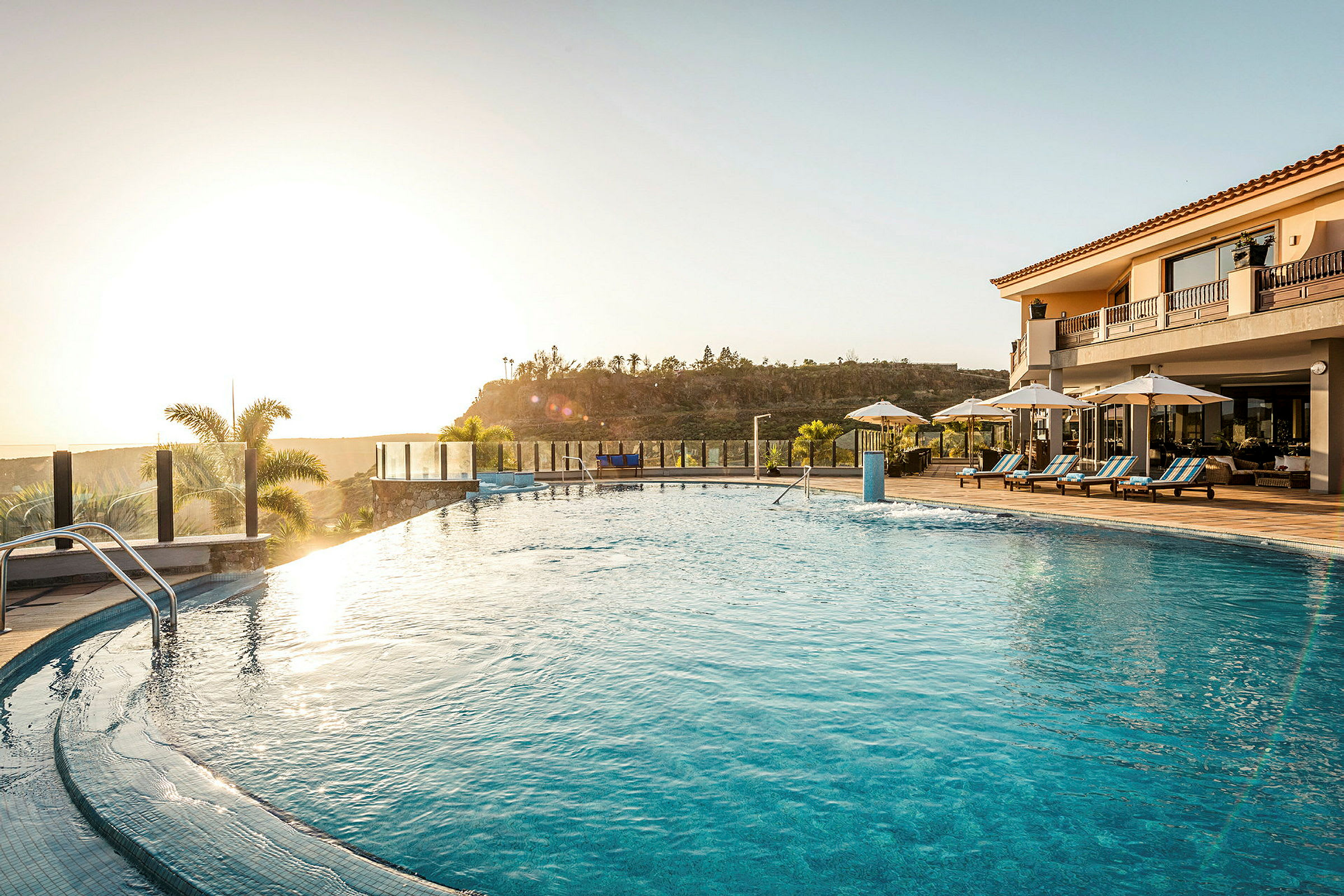 Udendørs swimmingpool ved solnedgang, omgivet af solsenge og parasoller. (AI-genereret tekst)