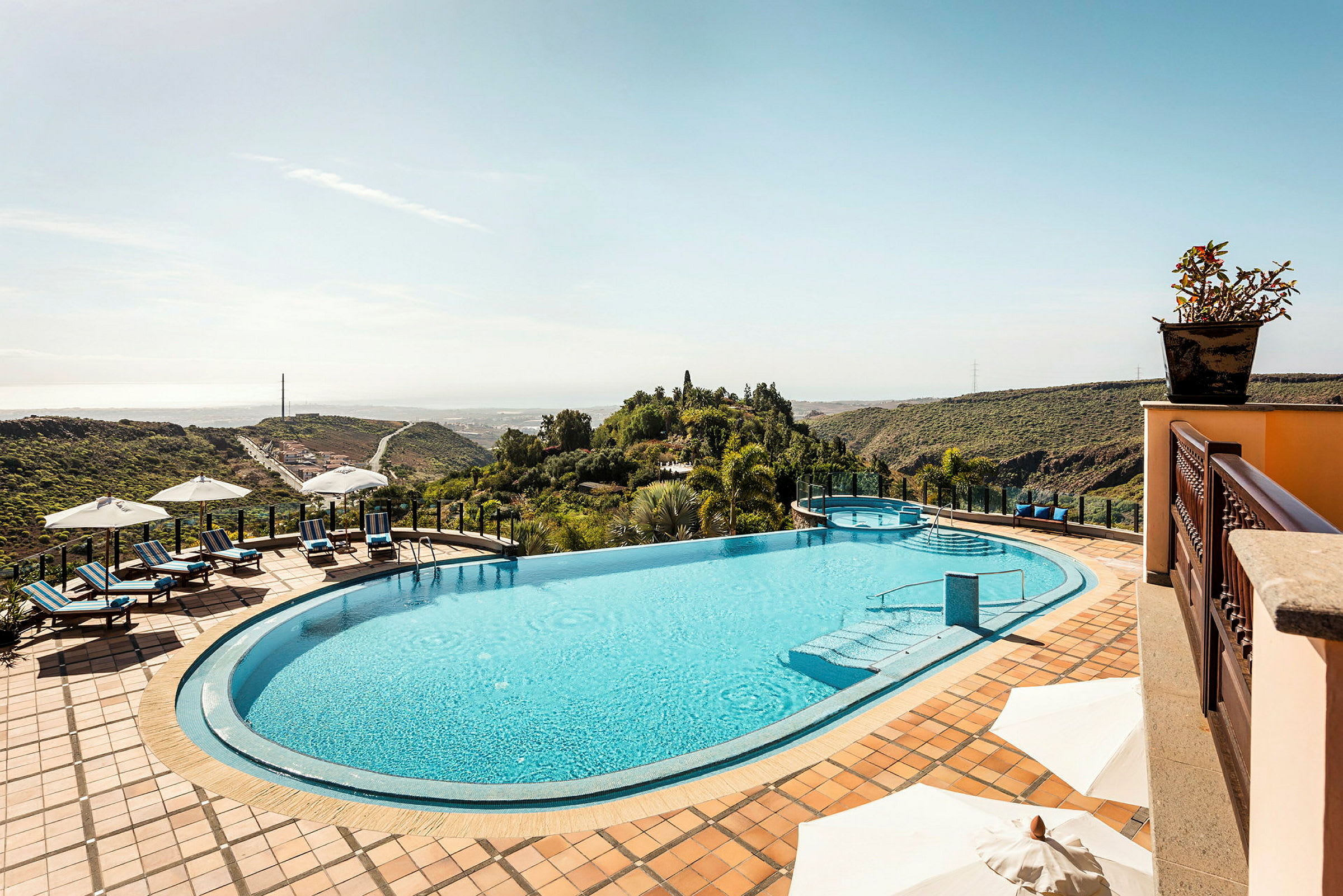 Udsigt over en infinitypool med liggestole og parasoller på en terrasse, omgivet af frodigt landskab. (AI-genereret tekst)