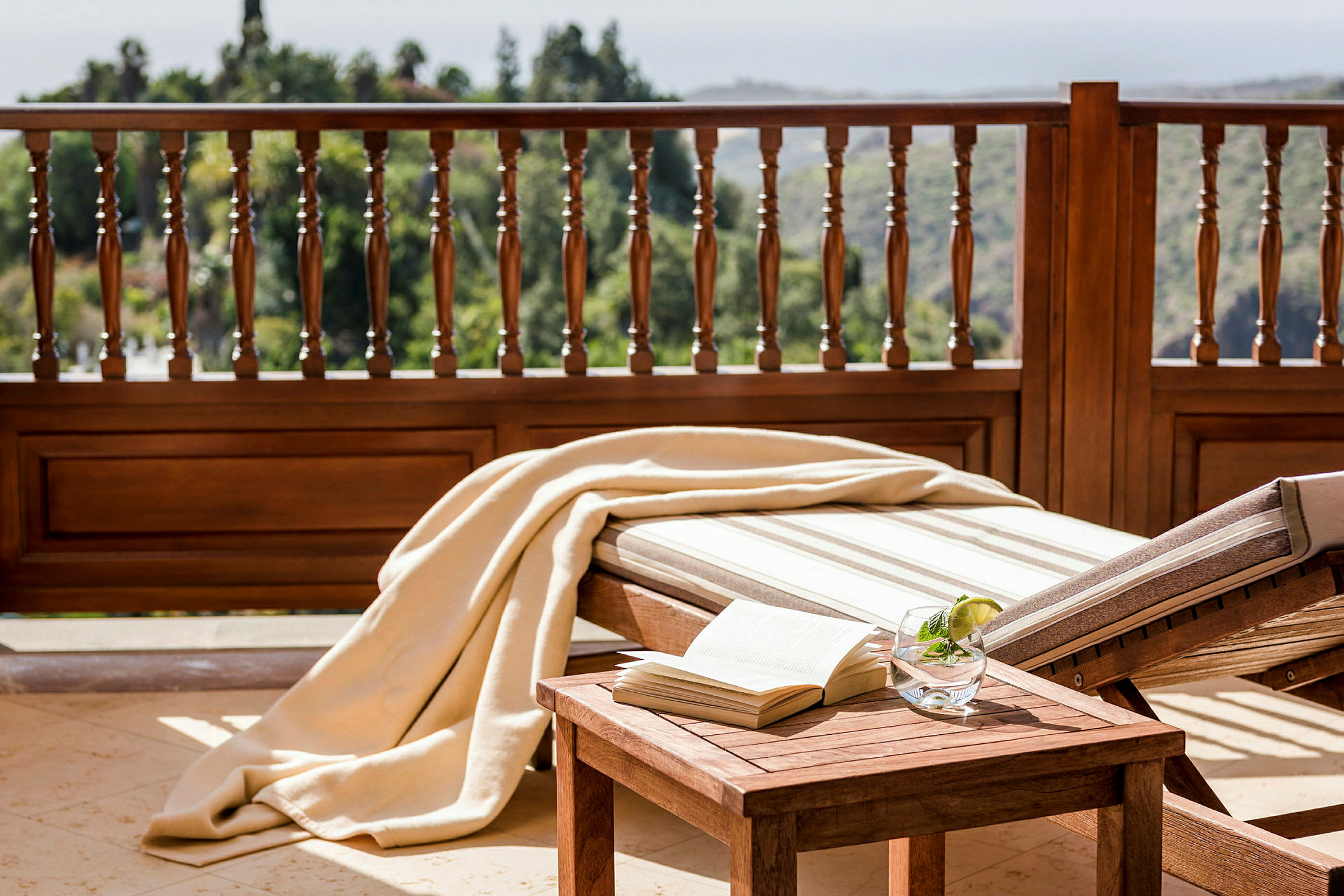 Liggestol og træbord med bog og drink, terrasse med træbalustrade og udsigt. (AI-genereret tekst)