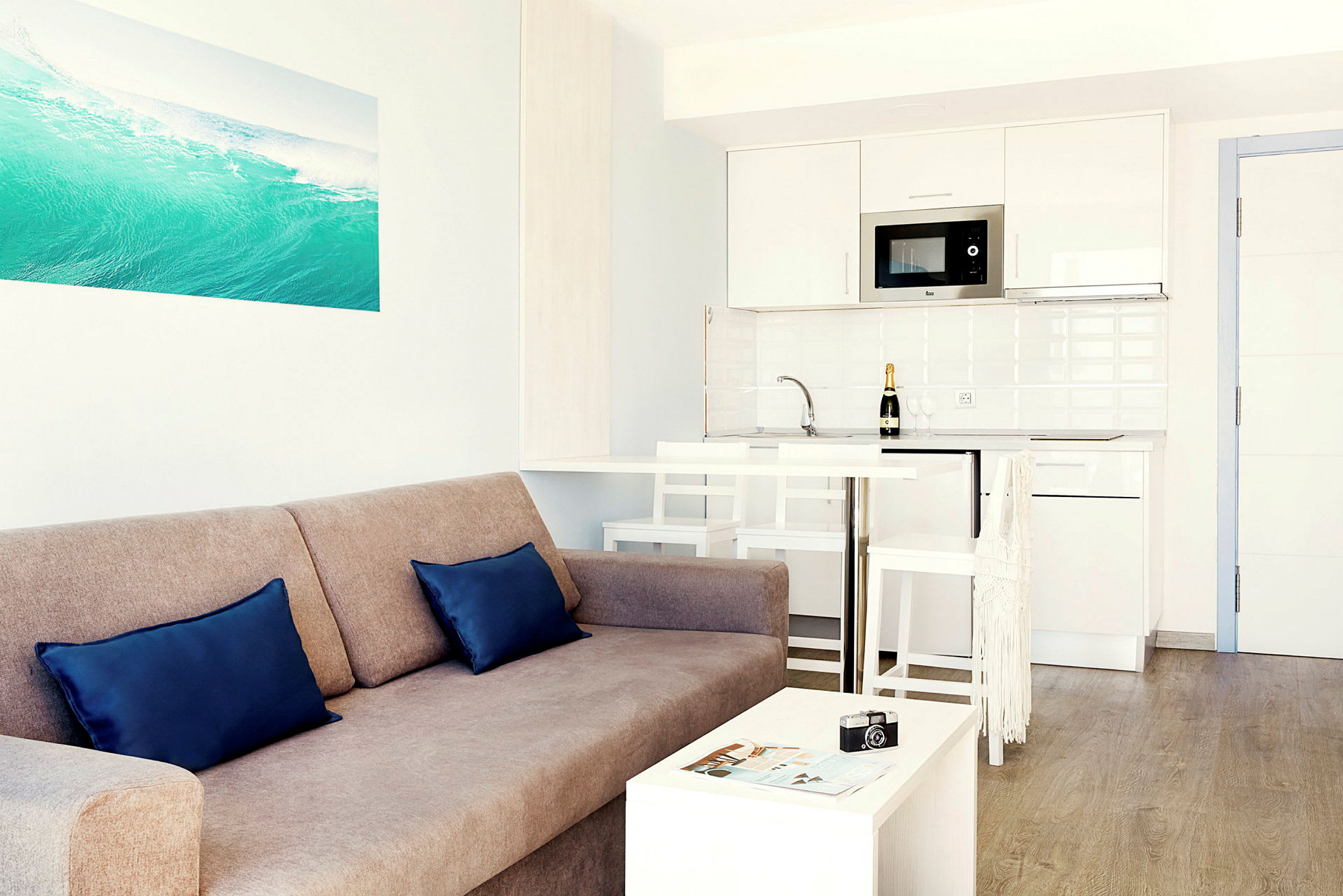 Et lyst og moderne opholdsrum med en sofa, to puder, et lille bord samt et køkken med hvidt interiør i baggrunden. (AI-genereret tekst)