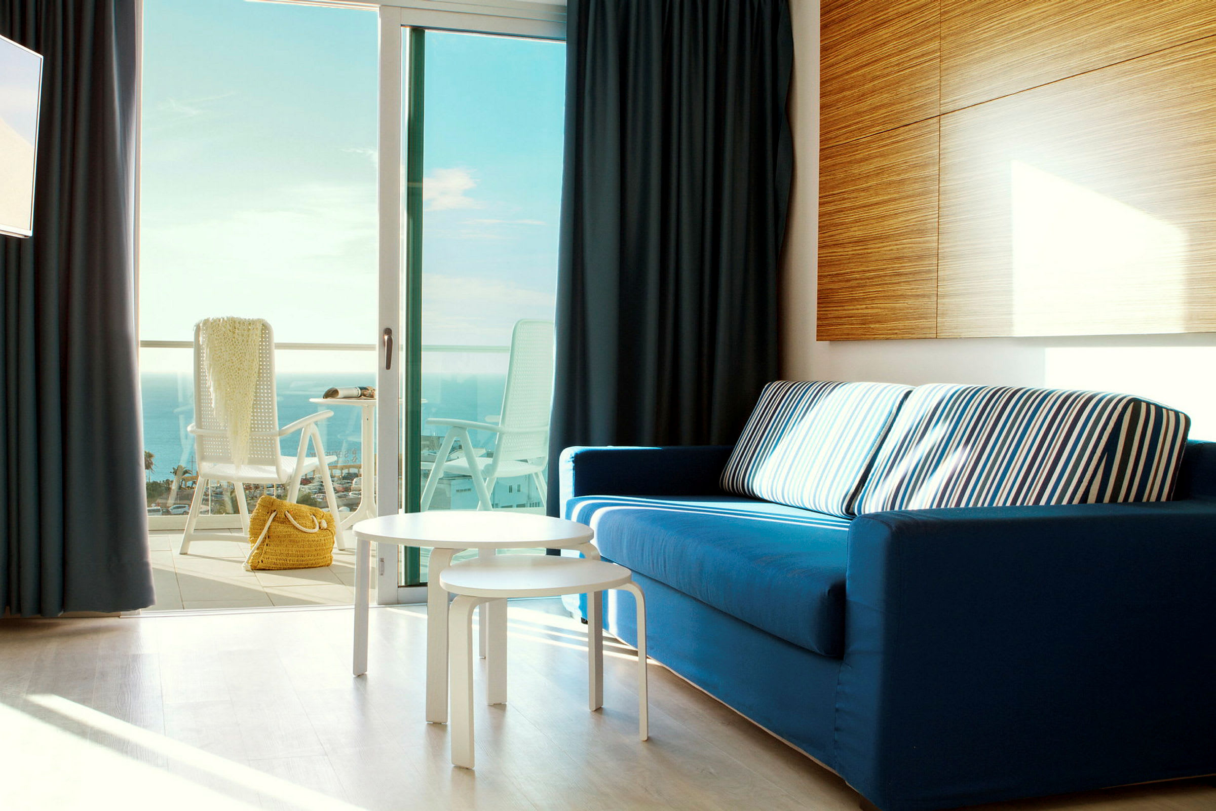 Et interiørbillede med en blå sofa, hvide borde og en åben altandør med udsigt over havet. (AI-genereret tekst)