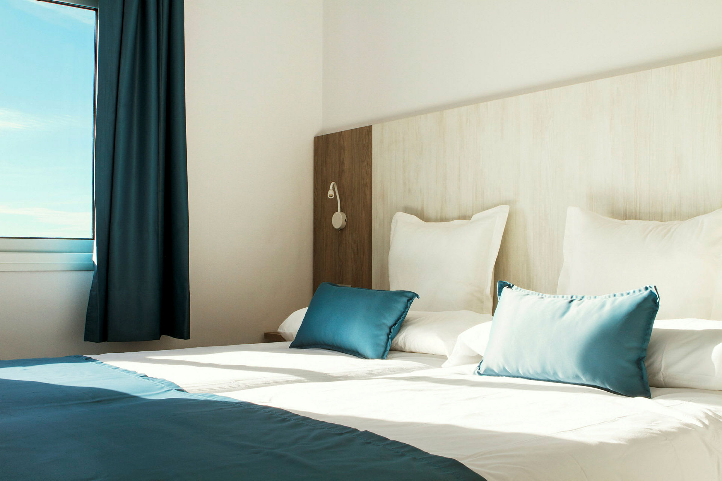 Interiør af et lyst hotelværelse med en opredt seng, hvide og blå puder samt et vindue, der lukker naturligt lys ind. (AI-genereret tekst)