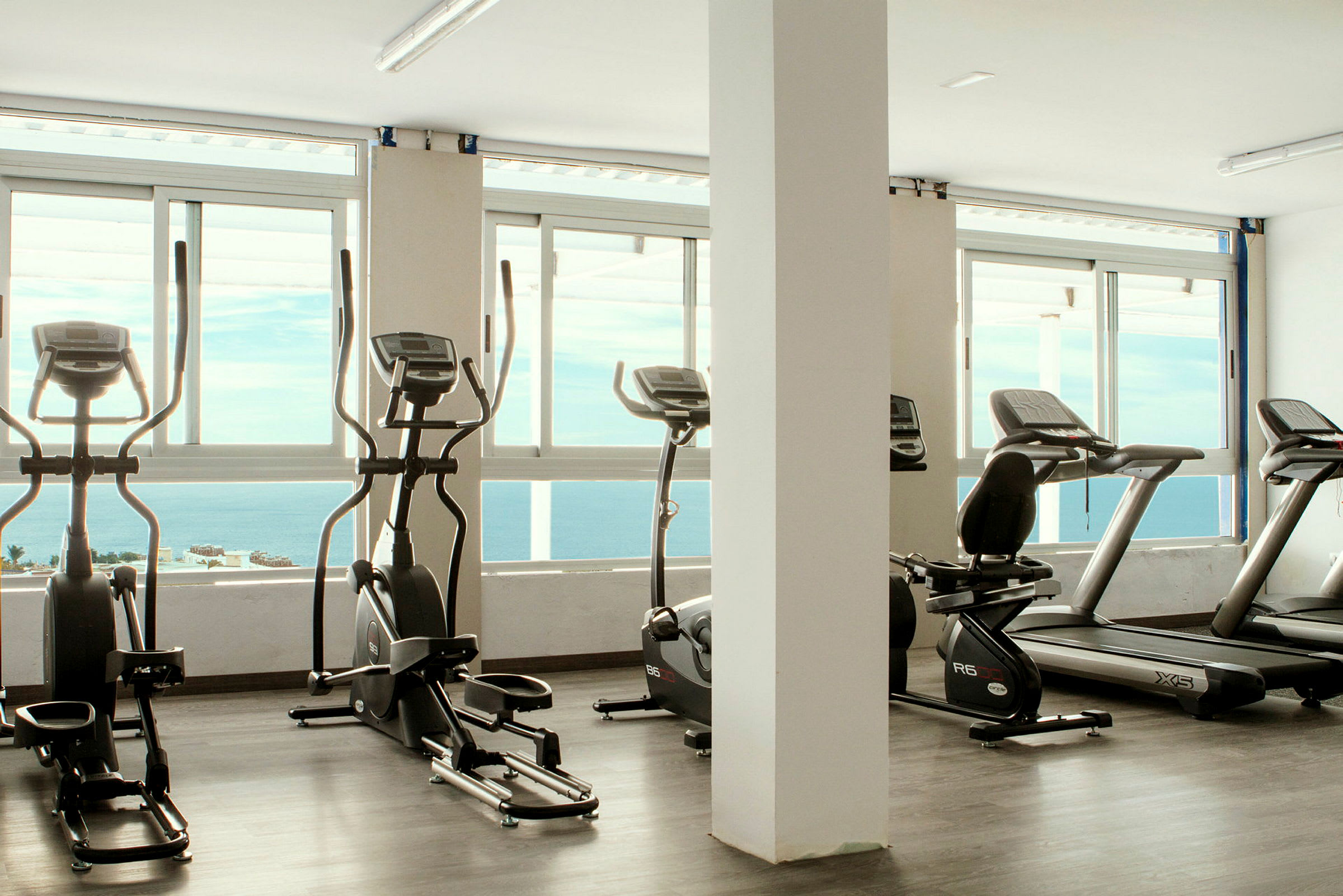 Fitnesscenter med træningsmaskiner foran vinduer med havudsigt. (AI-genereret tekst)