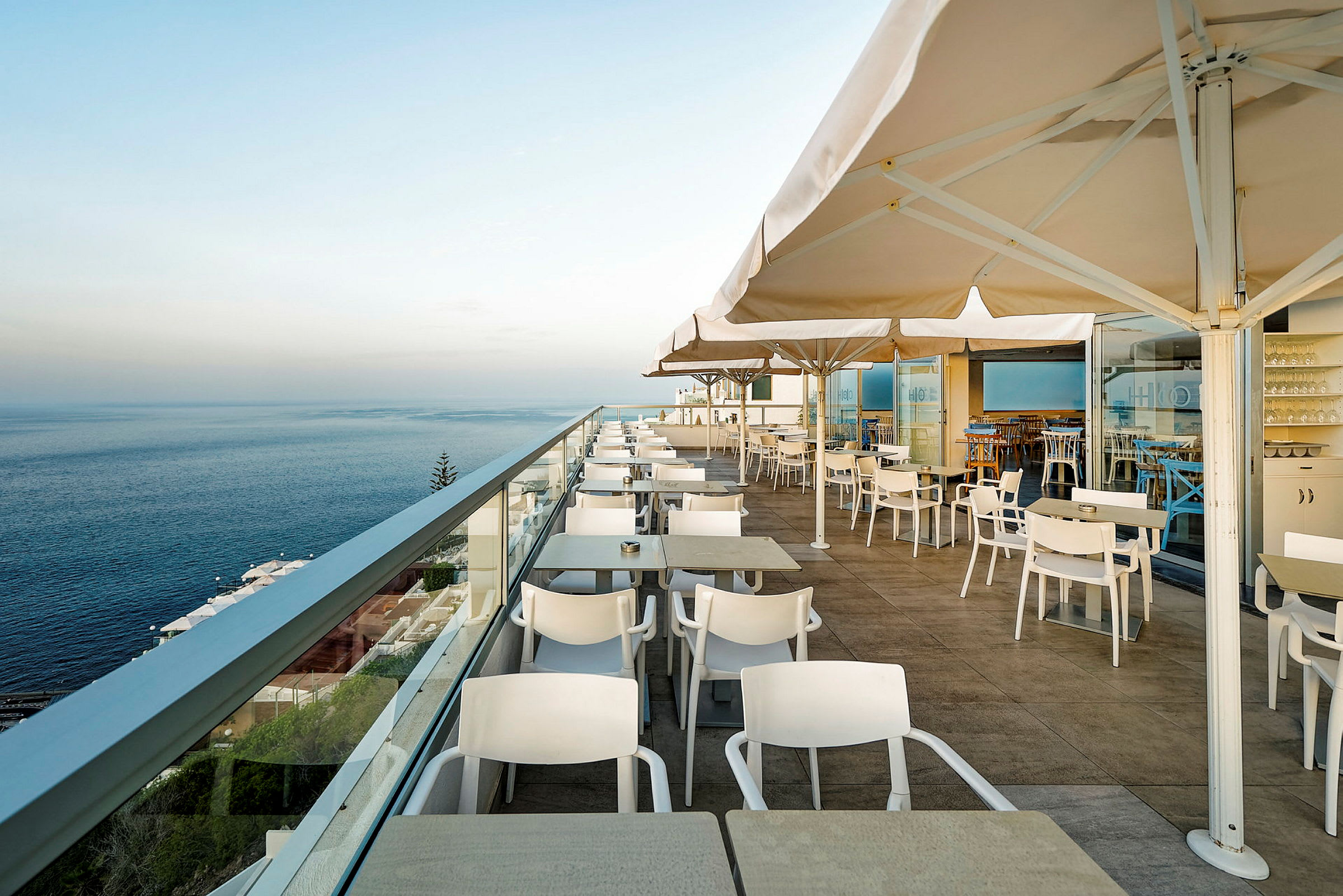 Billede af en udendørs terrasse med borde, stole og parasoller, med udsigt over havet. (AI-genereret tekst)