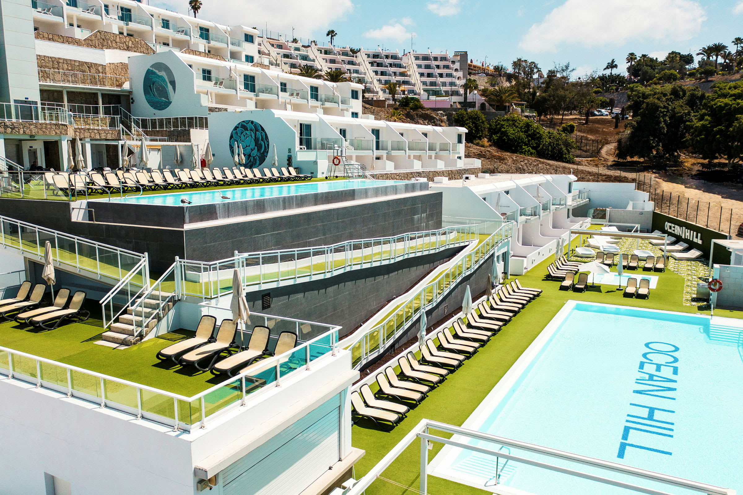 Billede af en resort med poolområder, liggestole og flere niveauer mod en skråning. (AI-genereret tekst)