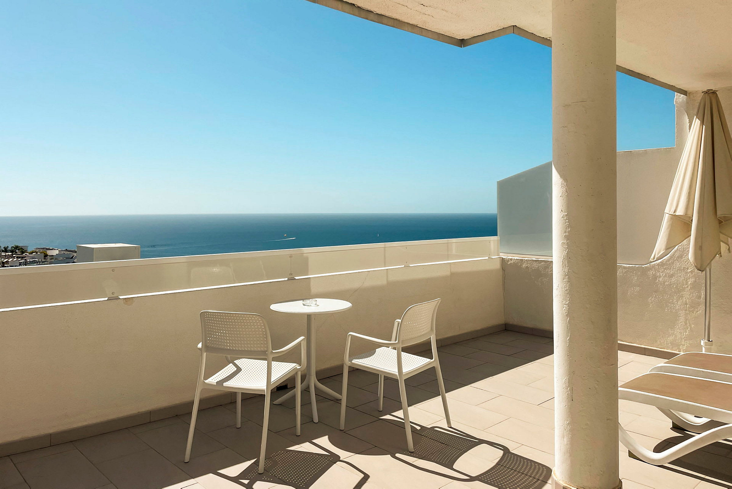 Terrasse med to hvide stole og et bord, havet ses i baggrunden. (AI-genereret tekst)