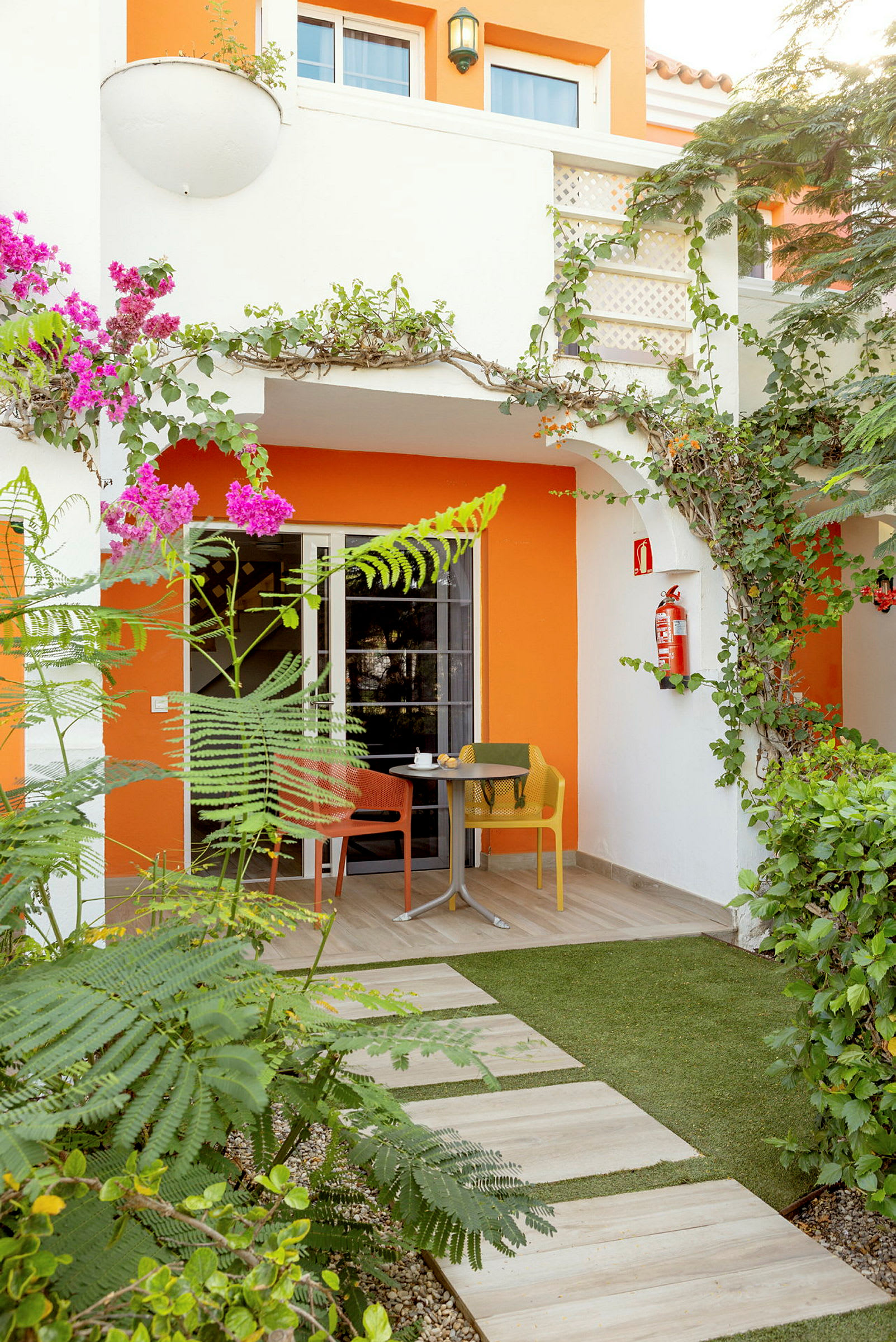 En terrasse med havemøbler omgivet af beplantning foran en bygning med orange og hvid facade. (AI-genereret tekst)