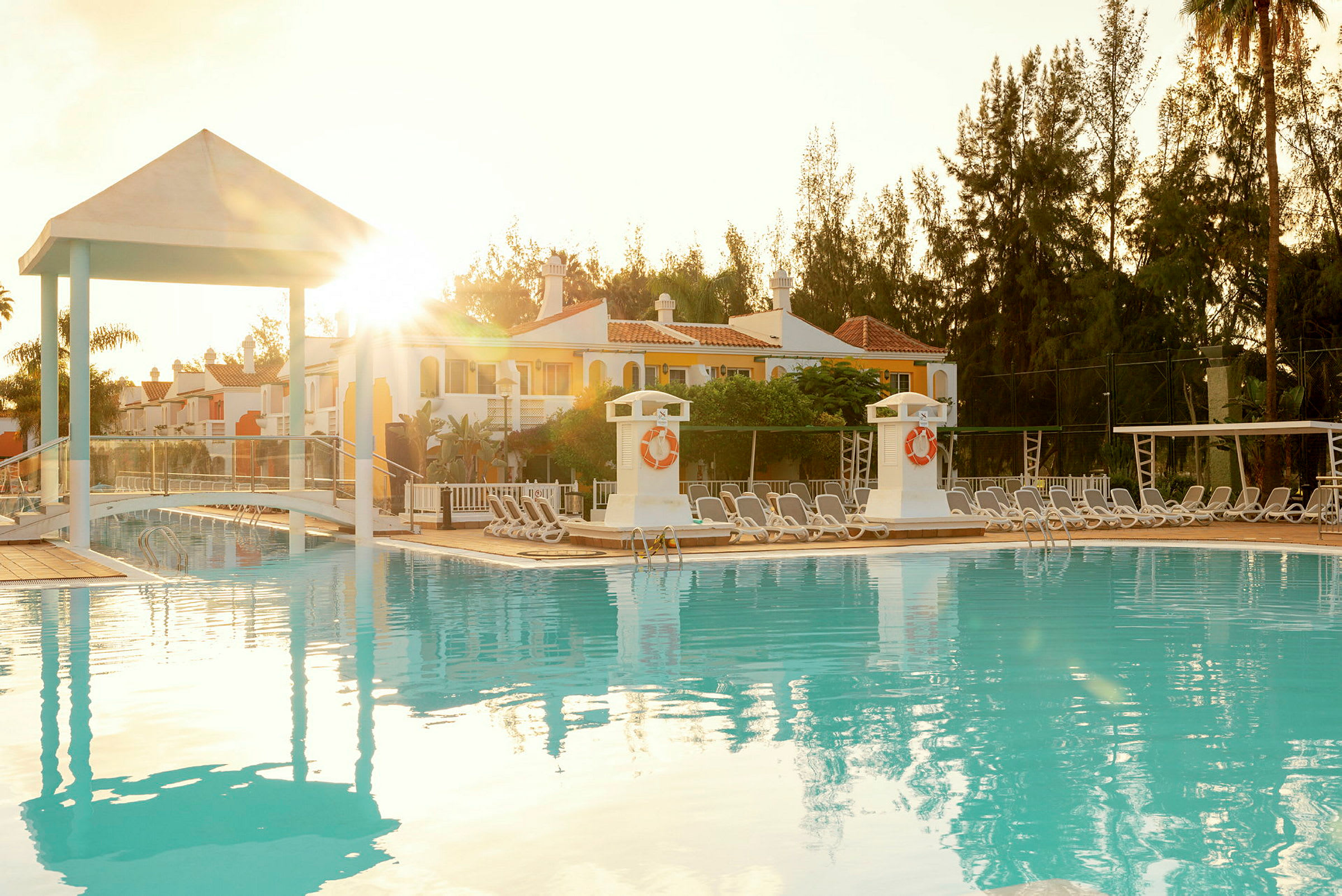 En udendørs swimmingpool med liggestole og bygninger i baggrunden, belyst af solen. (AI-genereret tekst)