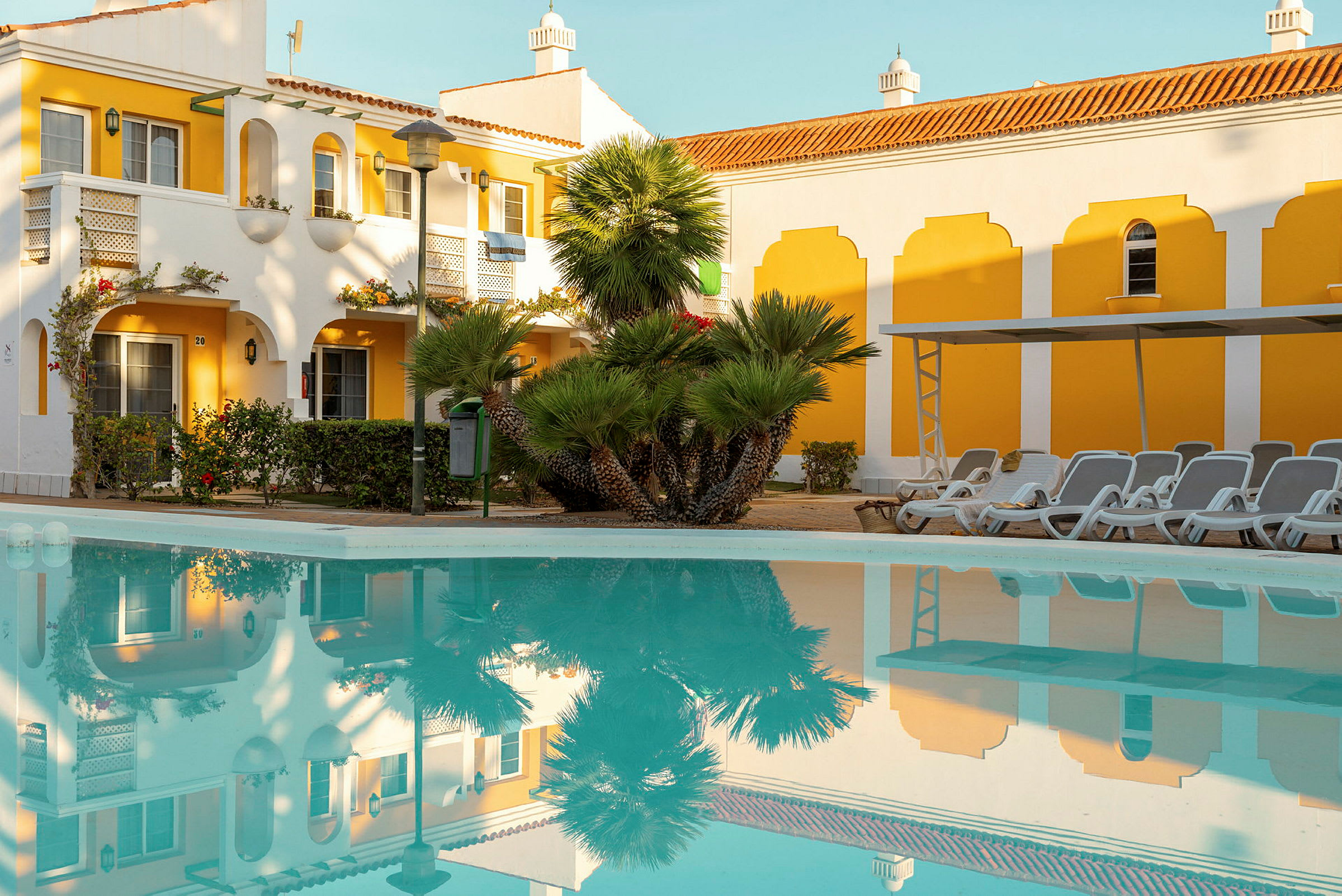 En gul og hvid bygning med buer ligger ved siden af en pool med refleksioner fra omgivelserne. Stole og parasoller er synlige ved poolen. (AI-genereret tekst)
