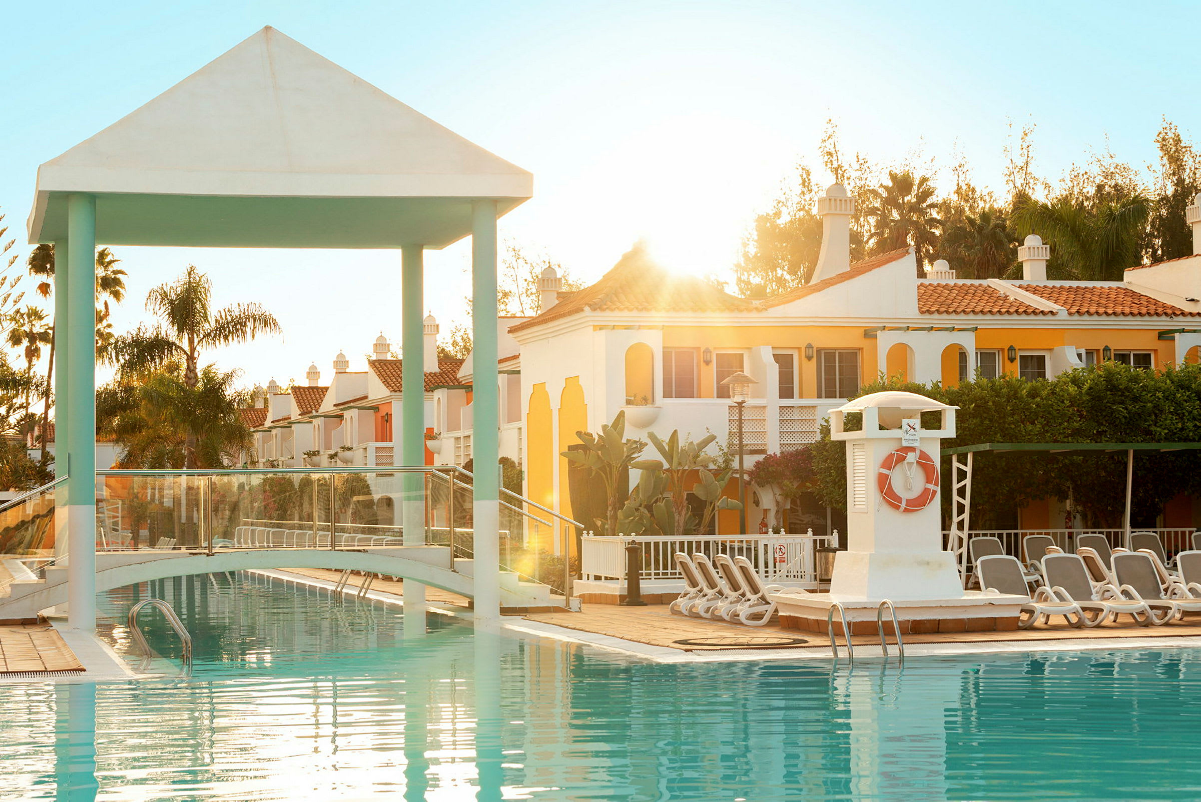 En udendørs pool med en lille bro, liggestole og farverige bygninger i baggrunden ved solnedgang. (AI-genereret tekst)