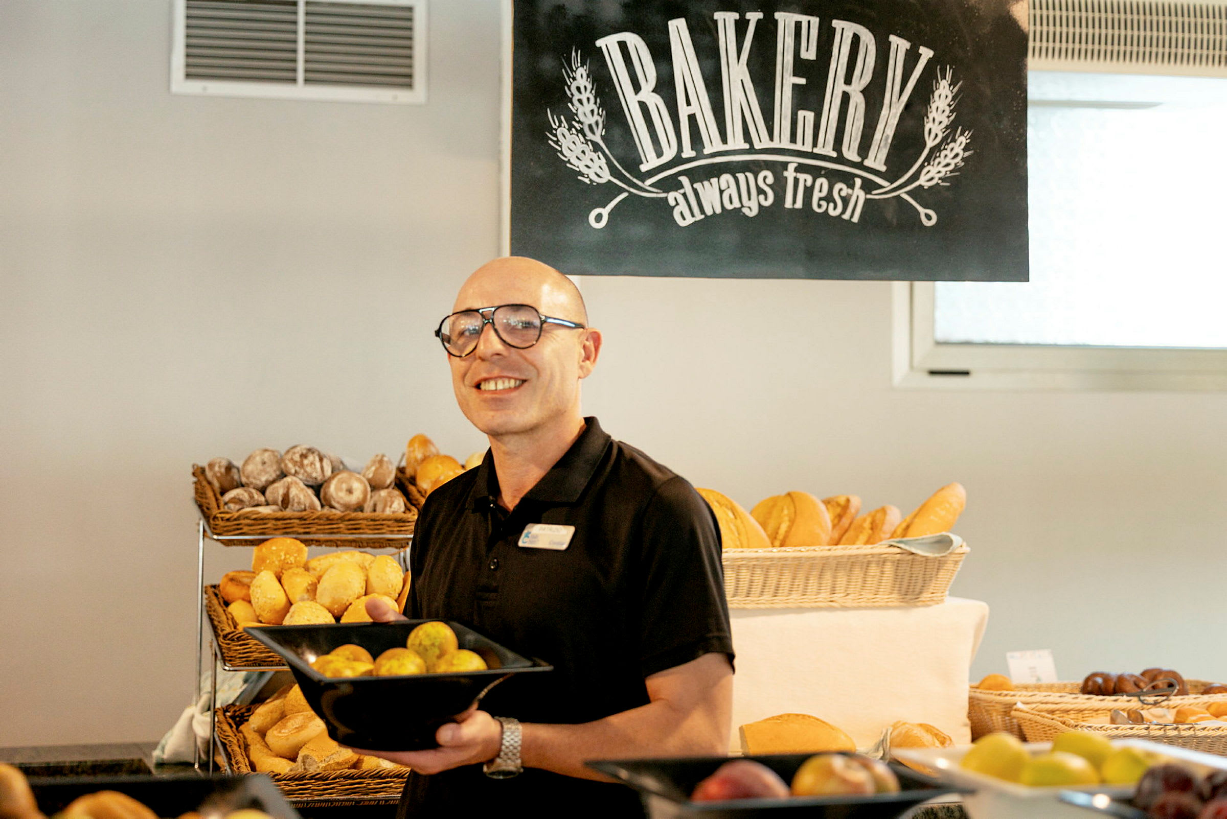 En person holder en sort skål fyldt med brød i et bageri, med andre bagværk og et skilt med teksten 'Bakery always fresh' synlige i baggrunden. (AI-genereret tekst)