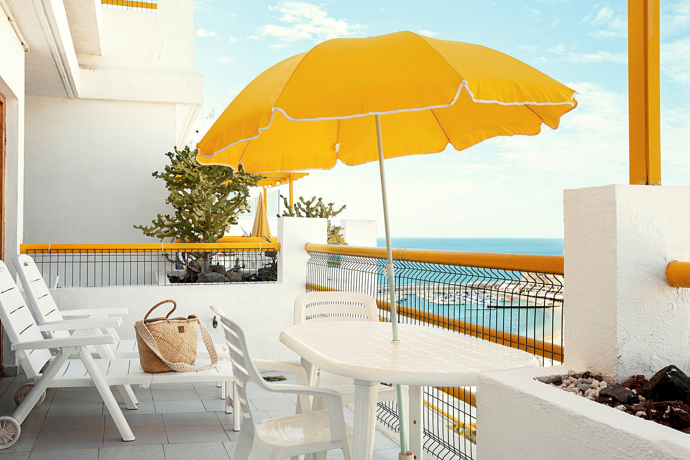 Hvid terrasse med stole, bord, gul parasol og havudsigt. (AI-genereret tekst)