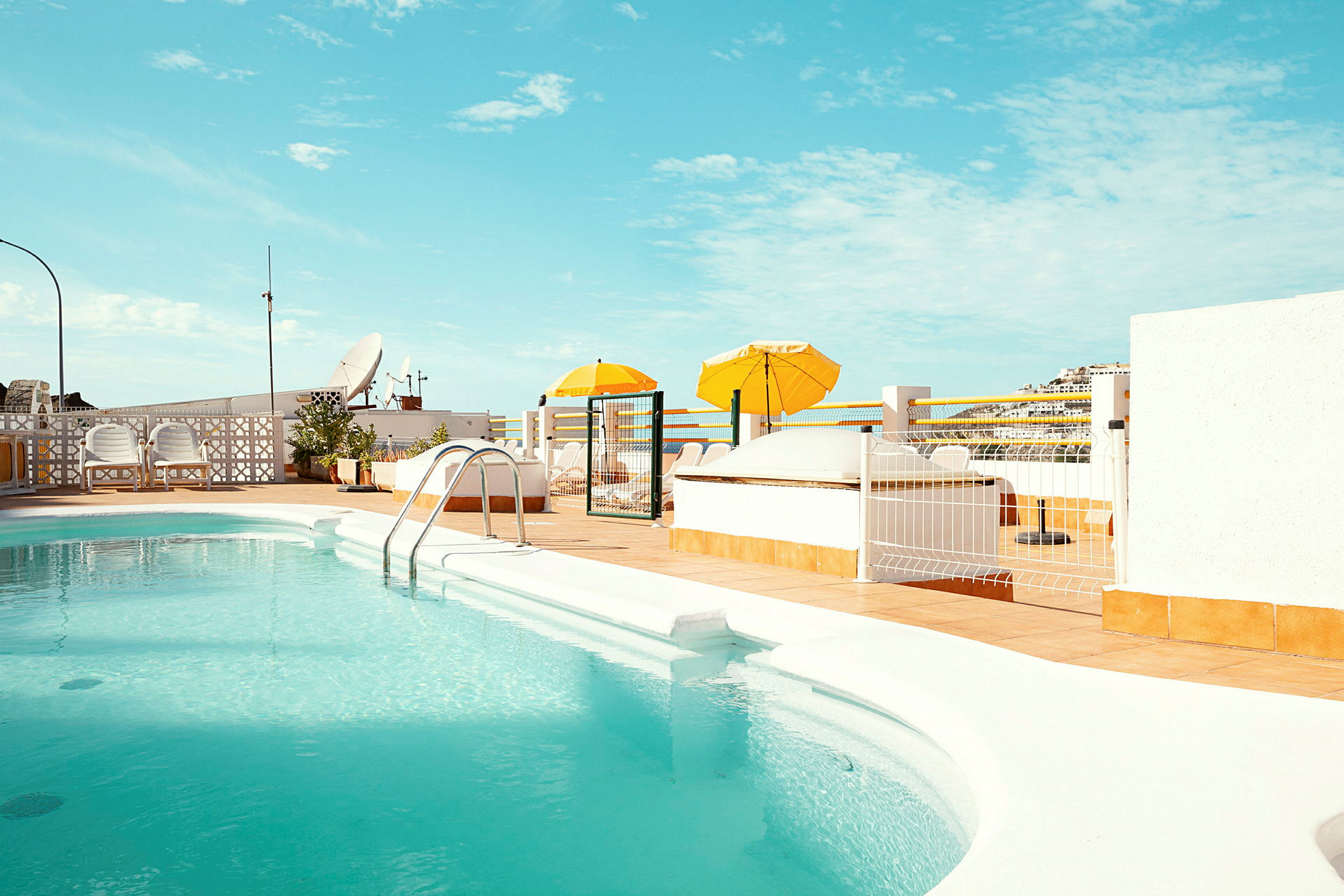 En udendørs pool med turkis vand omgivet af en solterrasse med gule parasoller. (AI-genereret tekst)