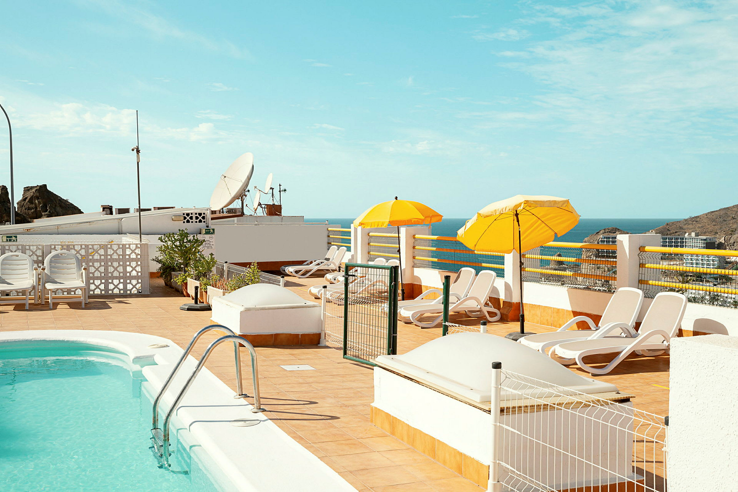 Tagterrasse med swimmingpool, liggestole og parasoller, udsigt til havet. (AI-genereret tekst)