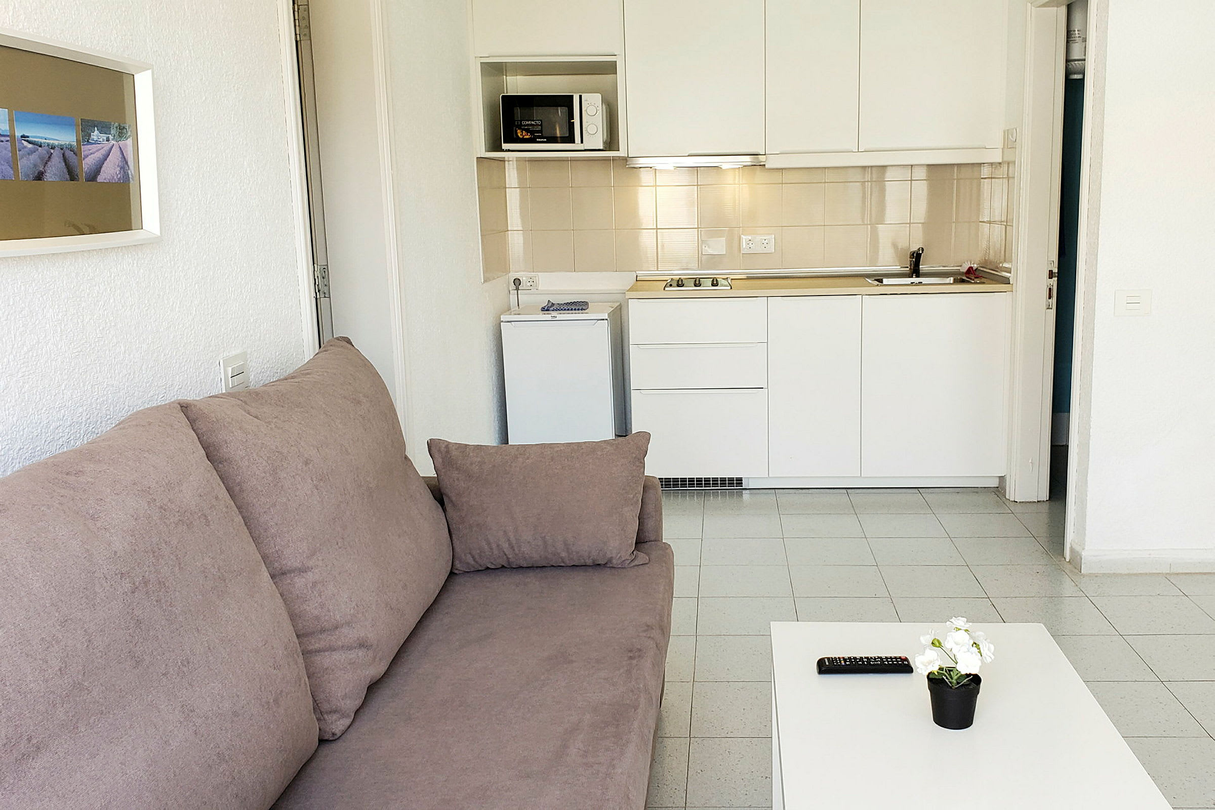 Et rum med en sofa, et bord med en blomsterpotte og fjernbetjening, samt et køkken med hvidt interiør. (AI-genereret tekst)